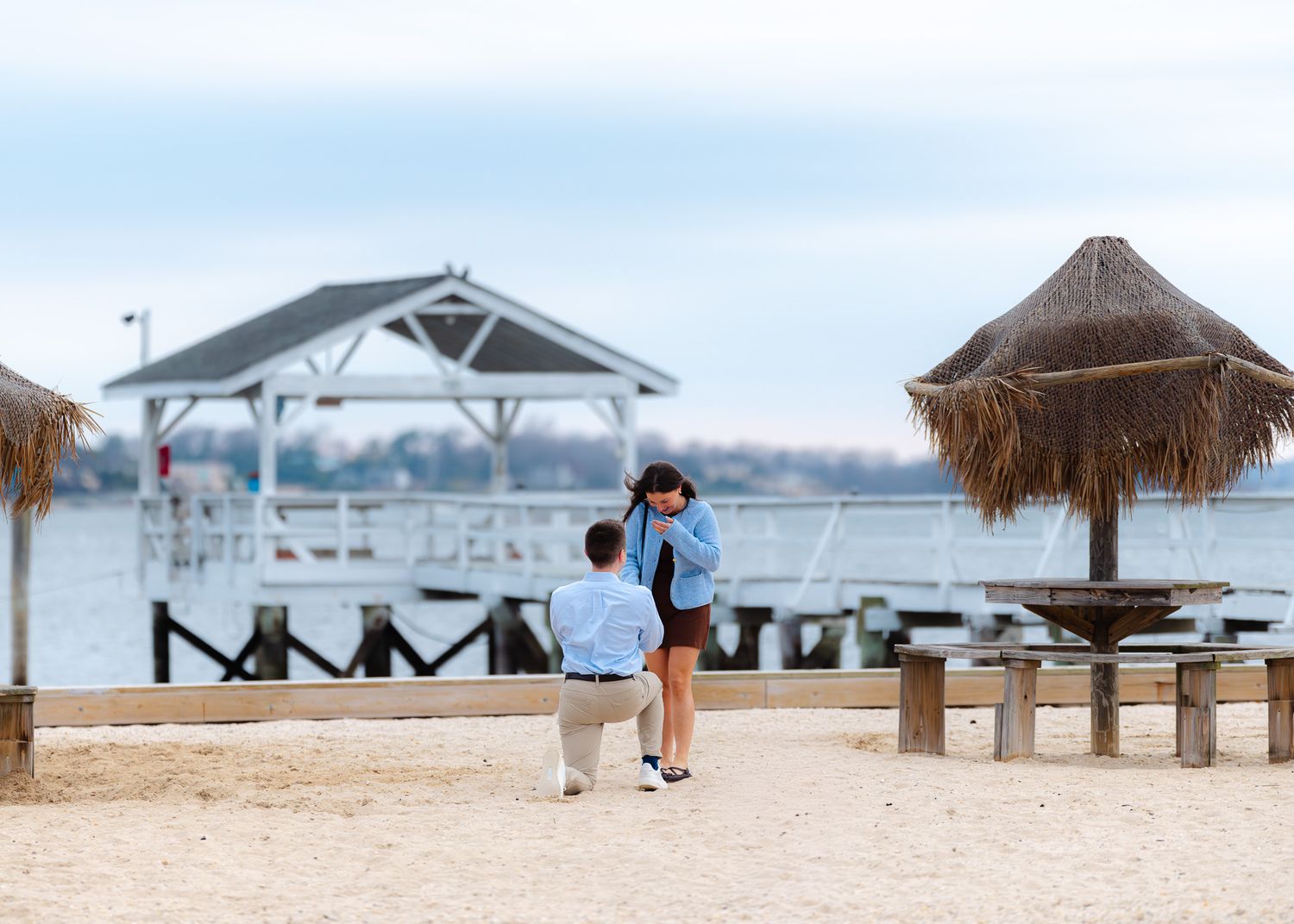 Man proposing on one knee to woman on sandy beach near waterfront dock and tiki huts.