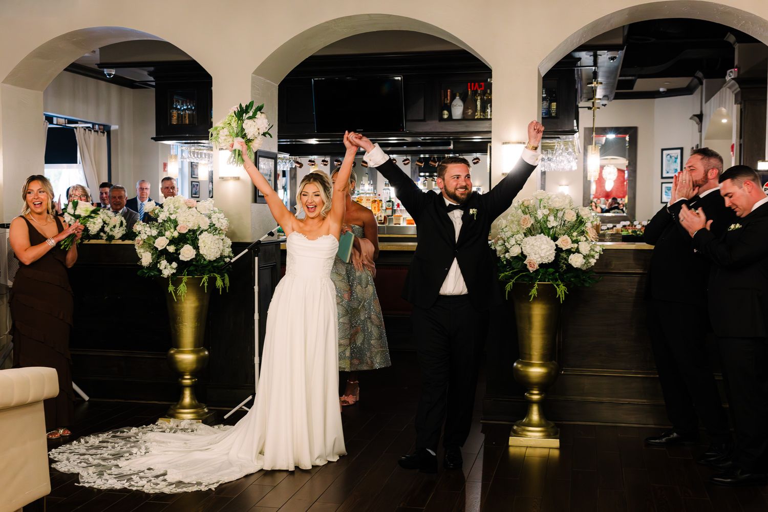 Newlywed couple raising hands in celebration at wedding reception, flanked by floral arrangements and applauding guests.