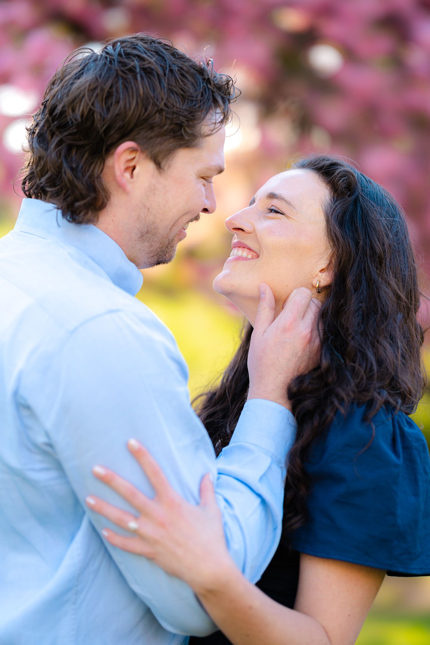 Couple nearly kissing and smiling near pink cherry blossom trees in a sunny park setting.