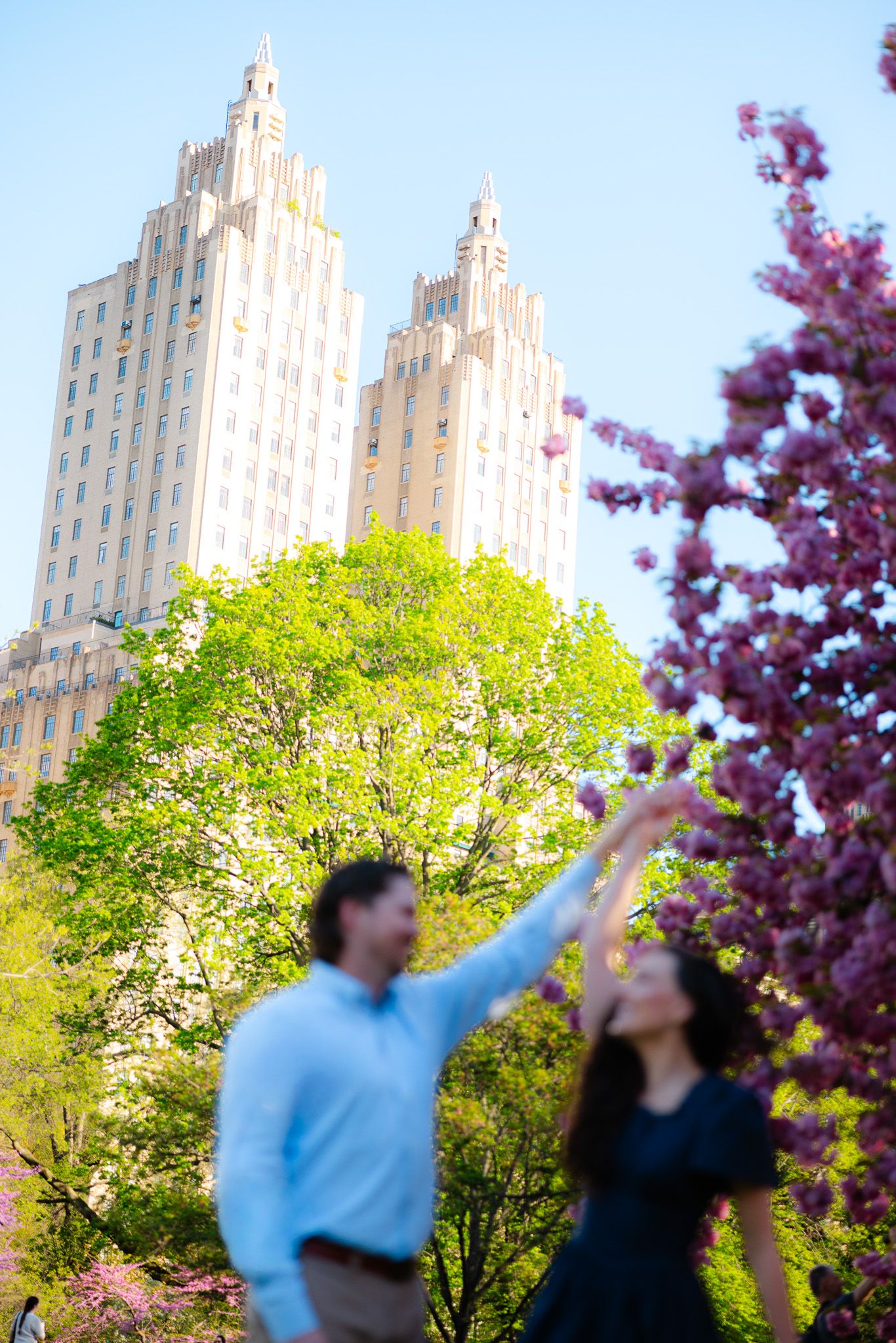 Couple reaching toward purple cherry blossoms with iconic NYC twin-towered building in background.