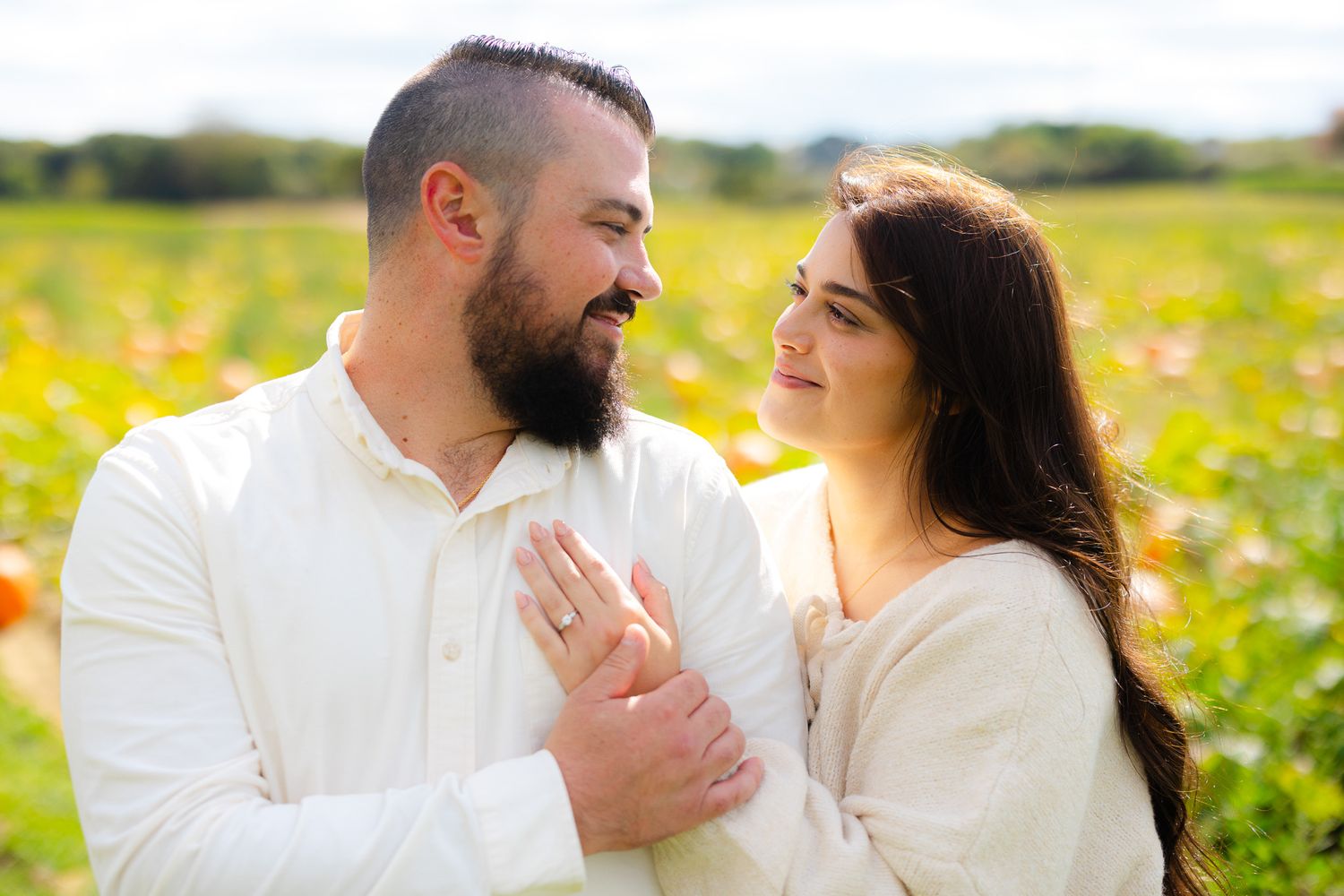 Happy engaged couple in white outfits smiling at each other in a colorful pumpkin patch on a sunny day.