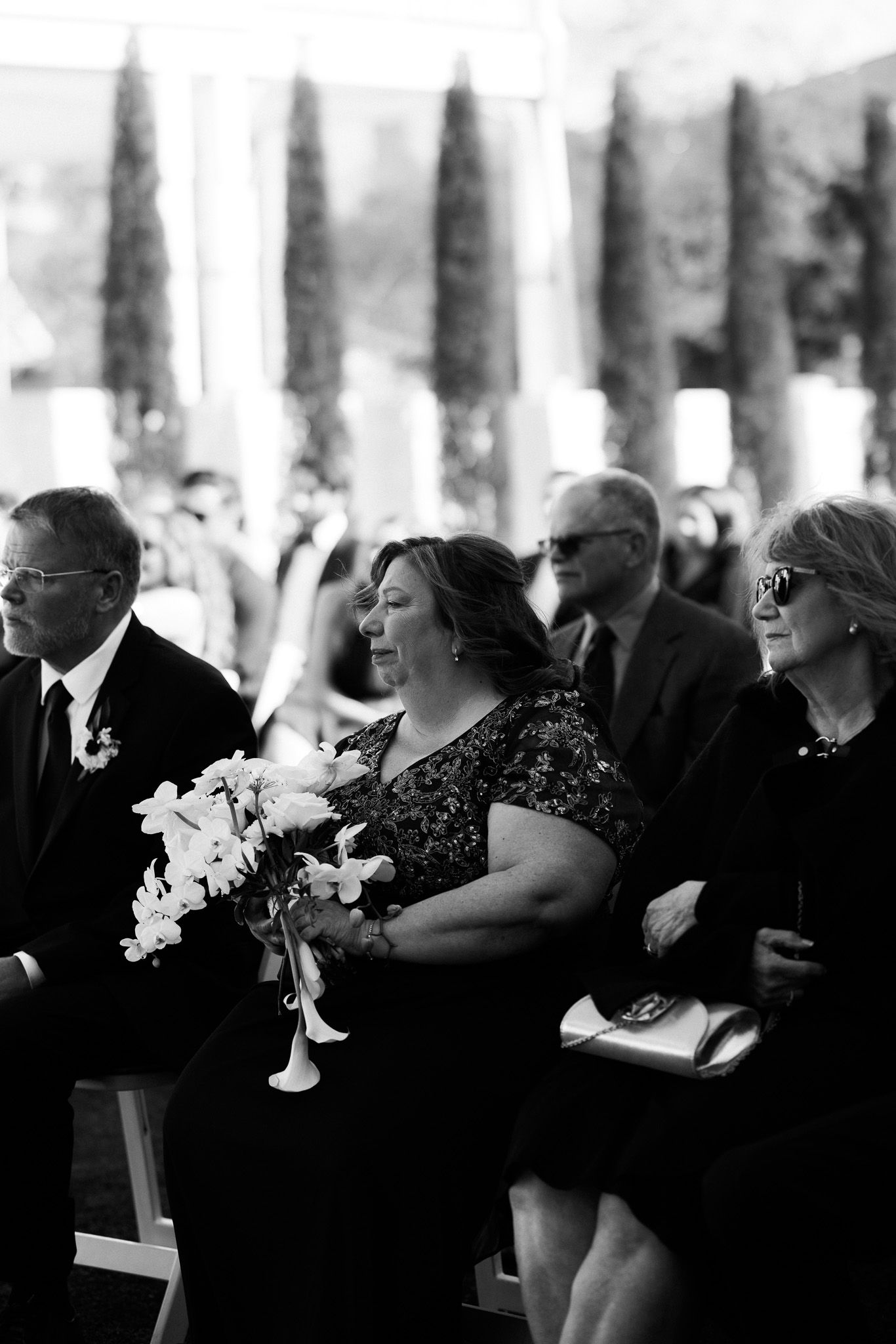 Wedding guests seated outdoors in black and white photo, woman holding white floral bouquet.