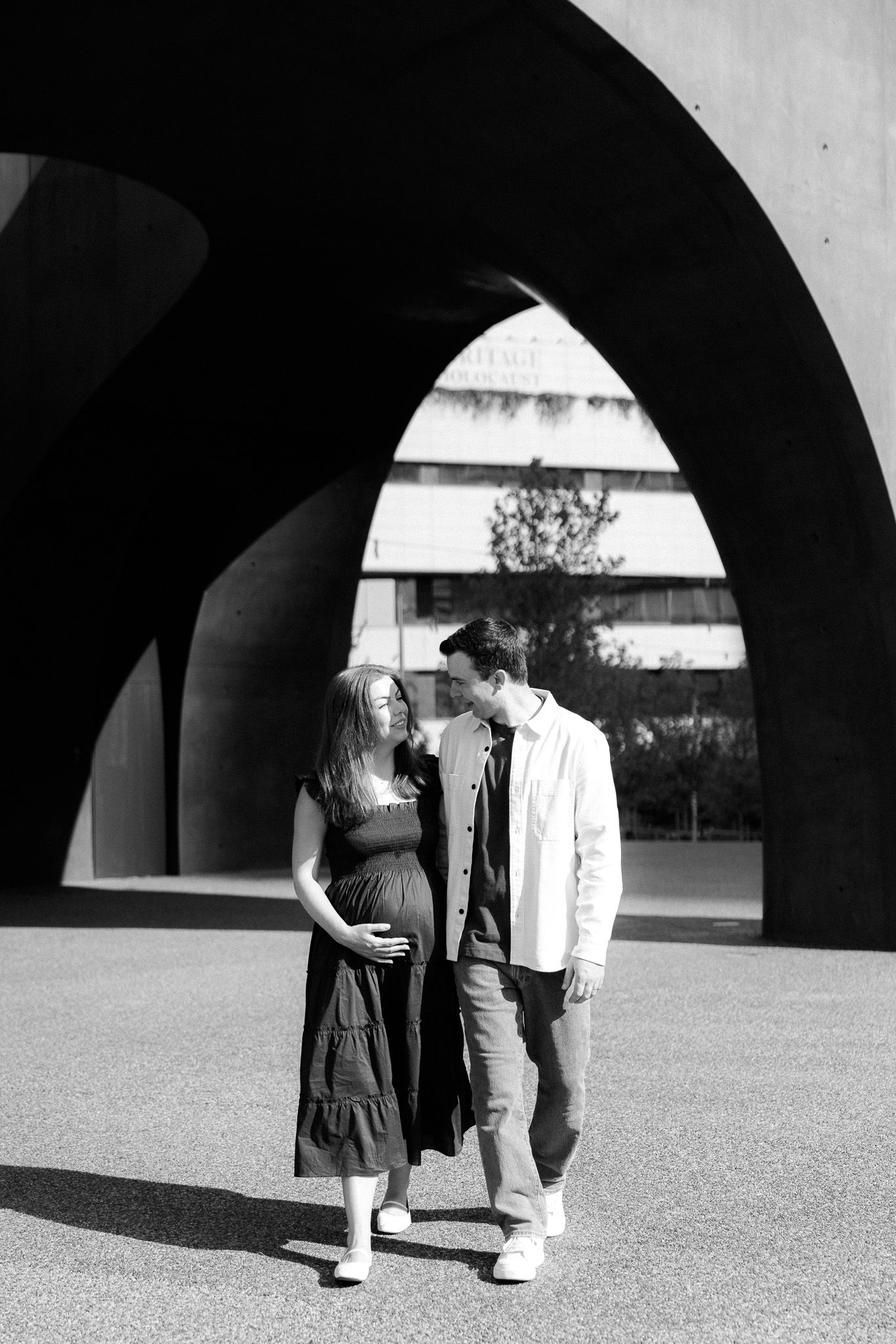 Black and white photo of a pregnant couple standing under concrete arches, smiling at each other outdoors.
