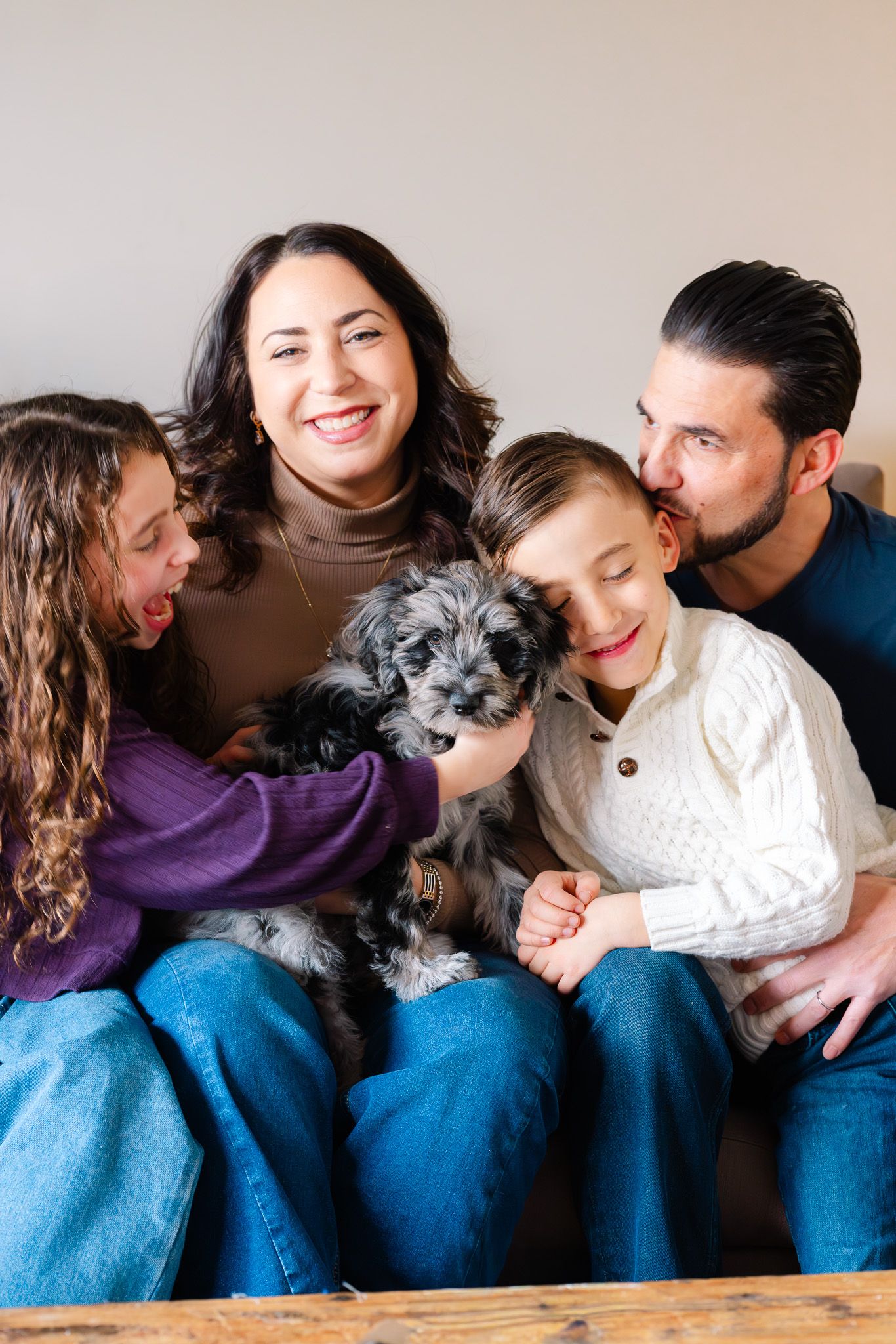 Happy family of four with a fluffy black and white puppy sitting together on a couch, smiling and laughing indoors.