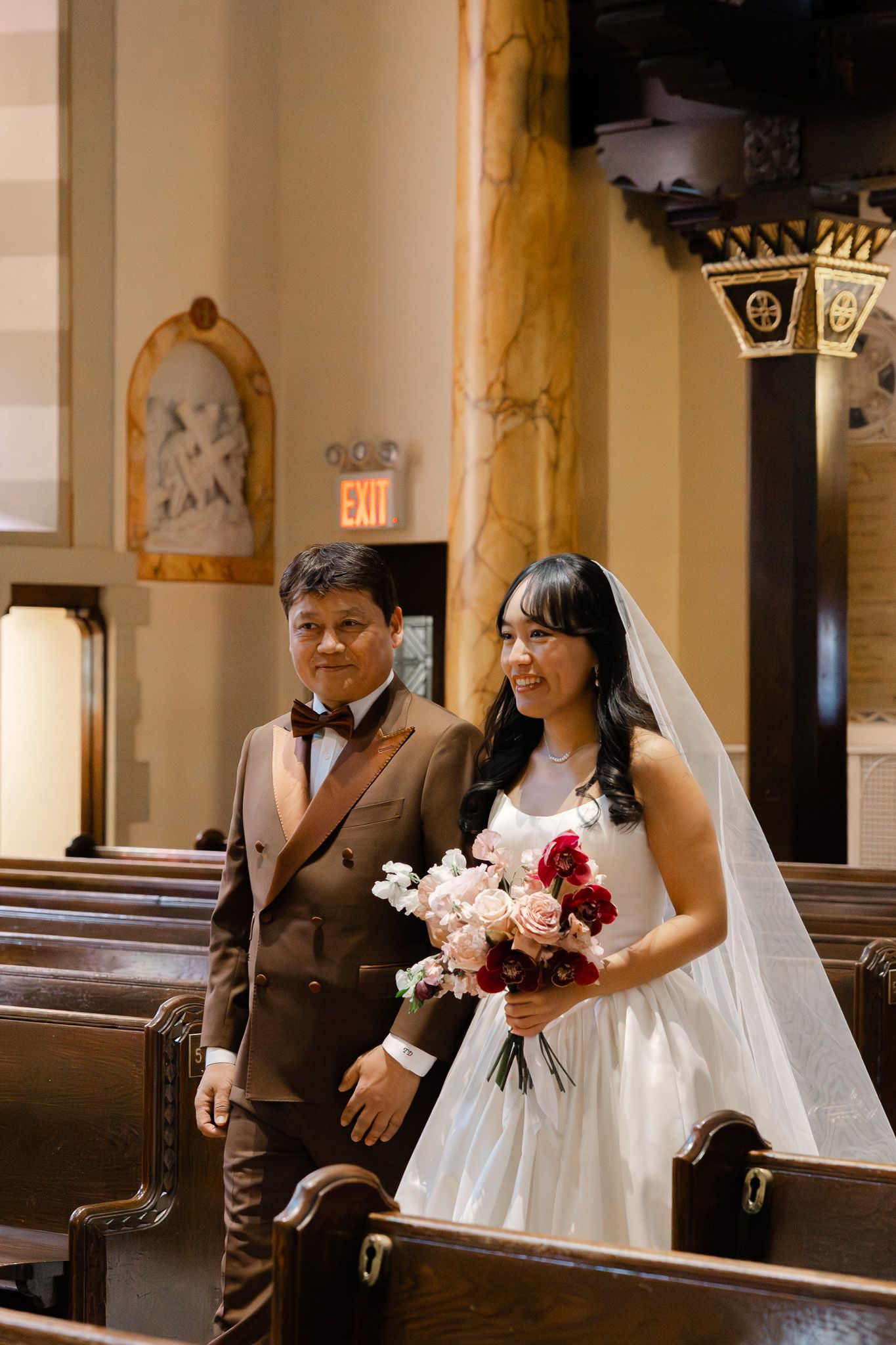 Bride in white gown and veil walks down church aisle with man in brown suit holding red and pink bouquet.