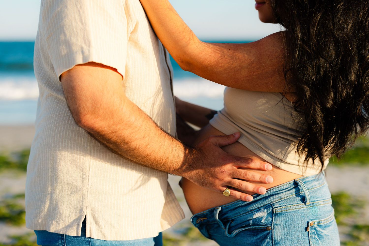 Couple embracing on a beach, man in white shirt holding pregnant woman in jeans near the ocean.