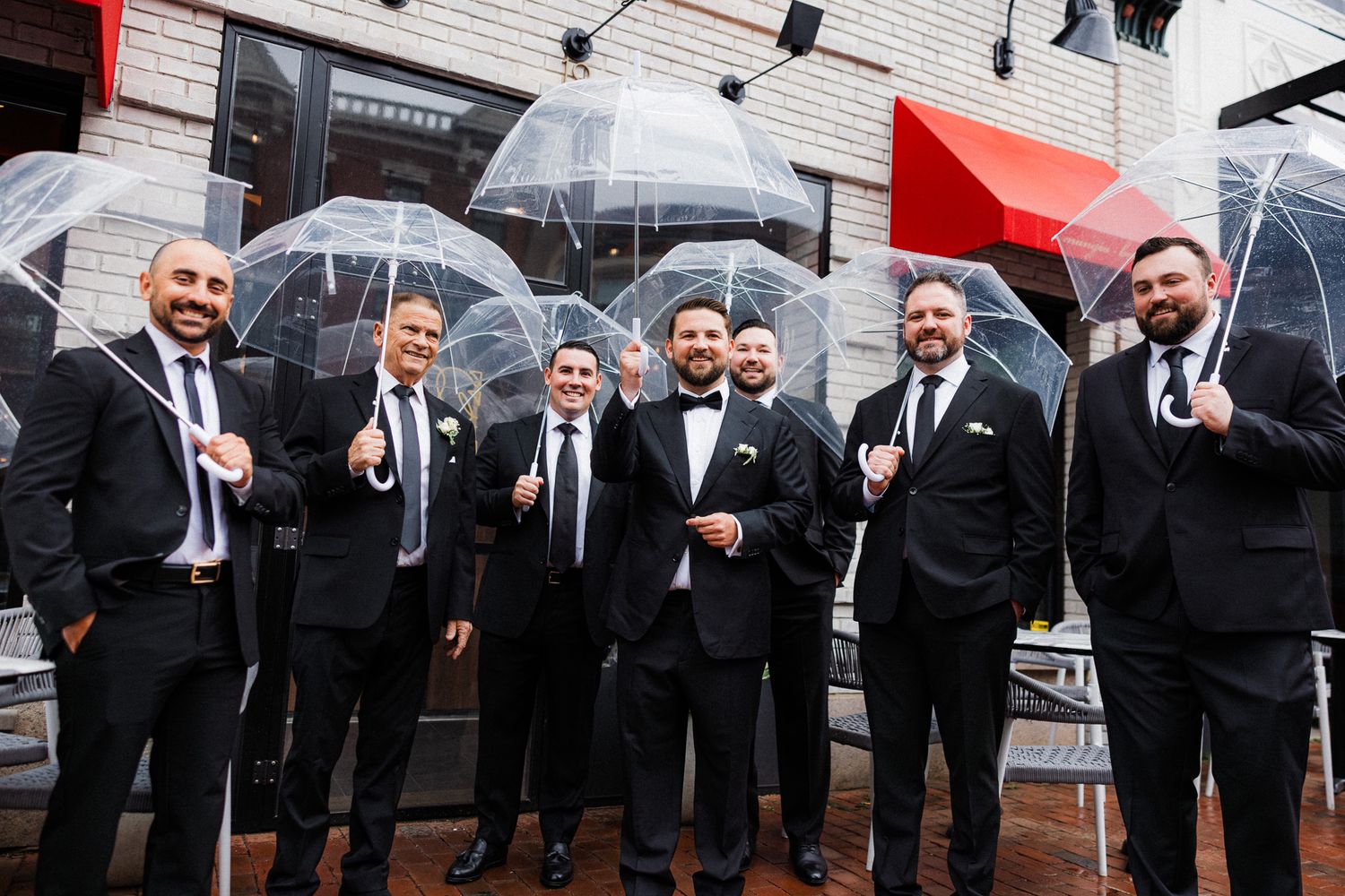 Seven groomsmen in black tuxedos holding clear bubble umbrellas posing outdoors on a rainy wedding day.