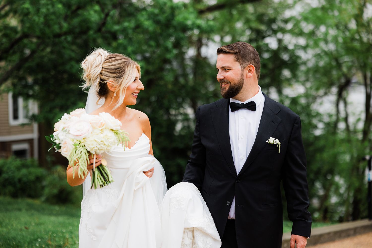 Bride and groom walking outdoors, she holds white bouquet in wedding dress, he wears black tuxedo with bow tie.