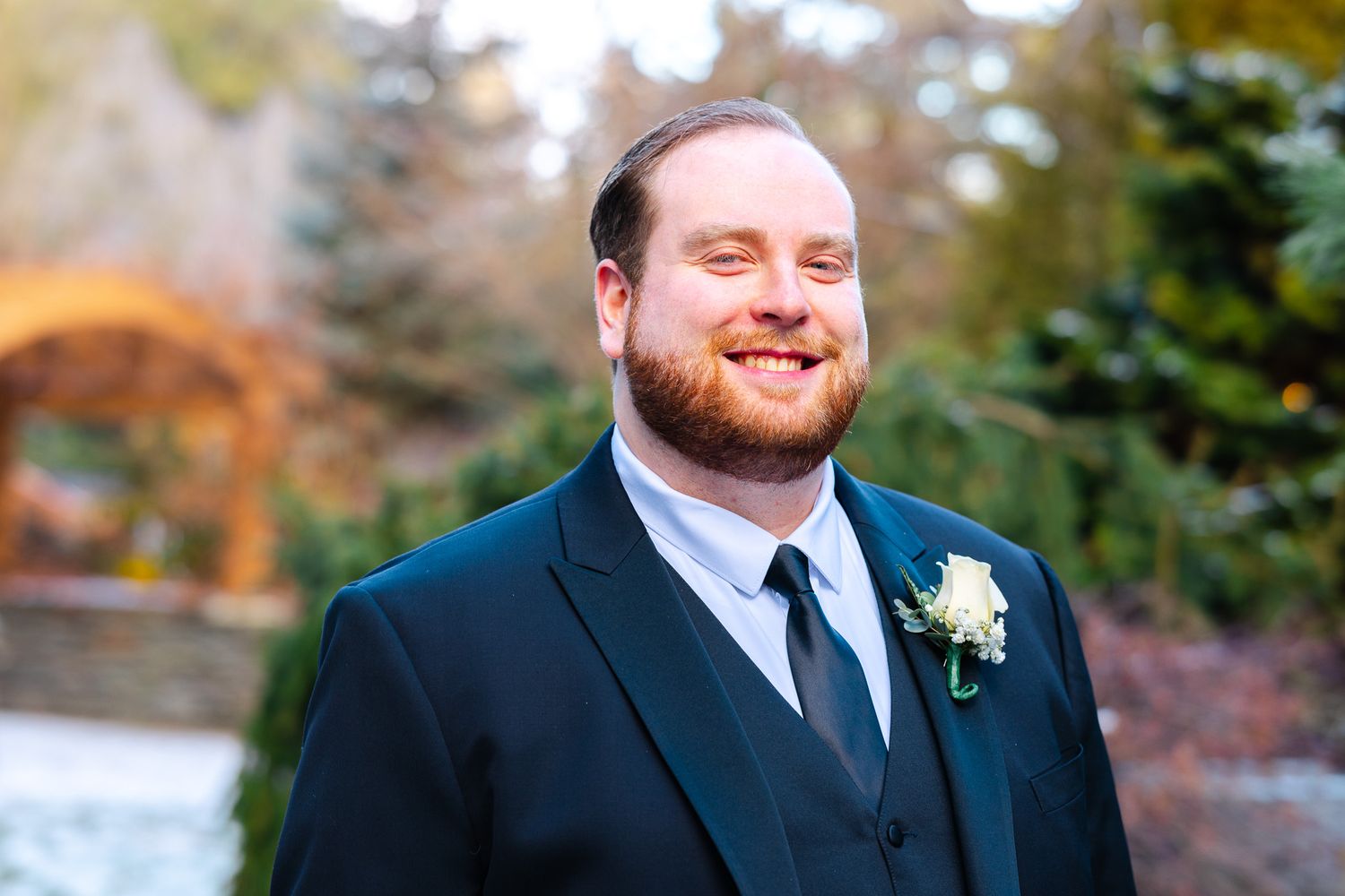 Smiling groom in dark suit with boutonniere poses outdoors at wedding venue with evergreen trees background.