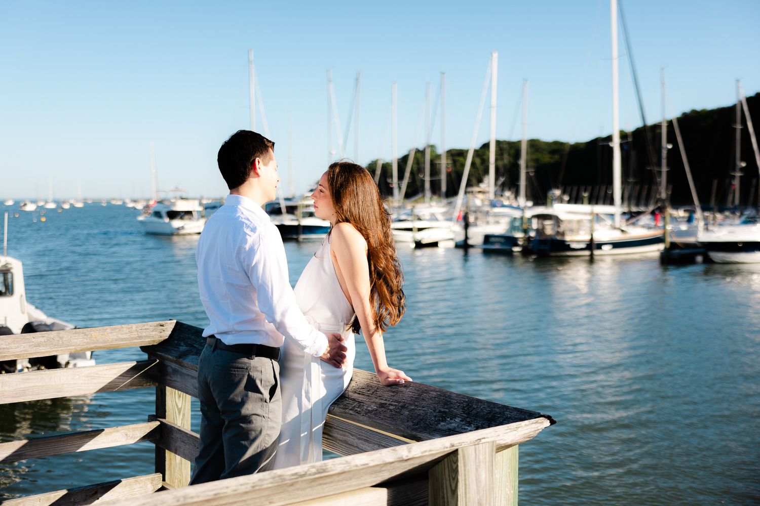 Couple standing on a wooden dock overlooking a marina with sailboats on a sunny day.