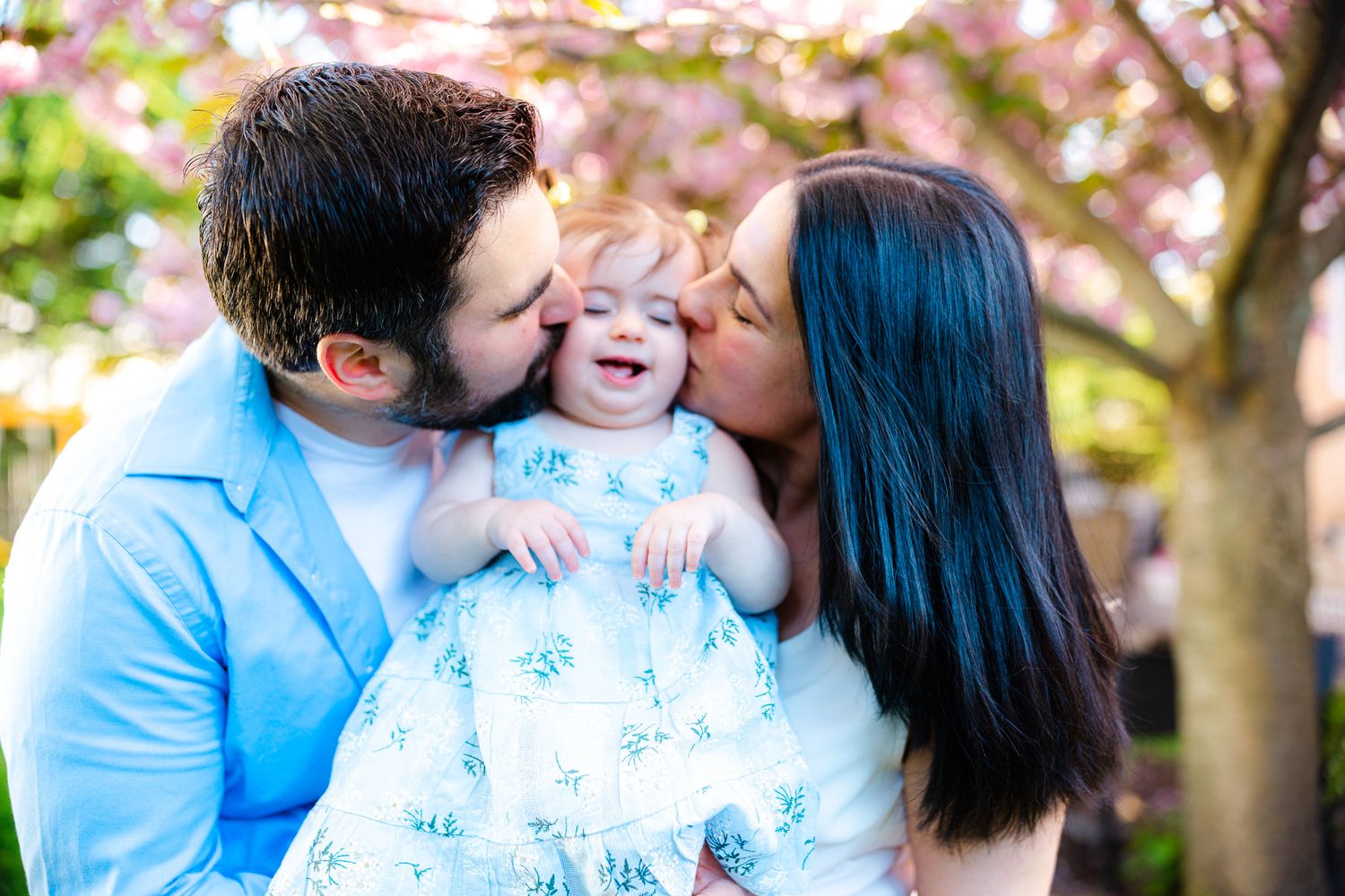 Parents kissing their smiling baby girl on cheeks outdoors with pink cherry blossoms in spring.