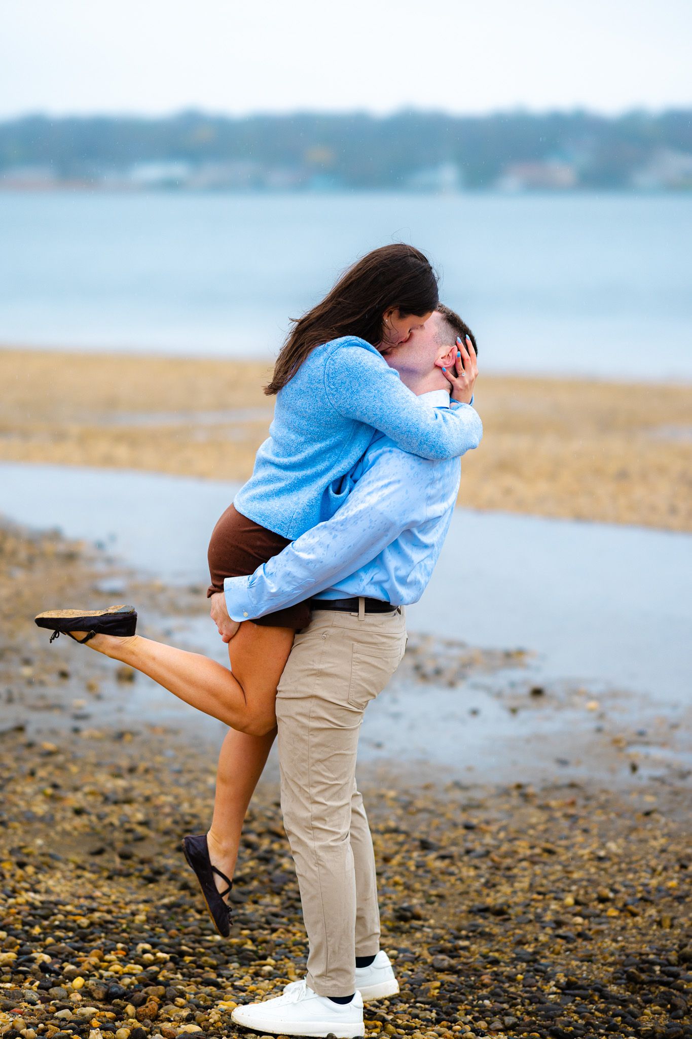 Couple sharing a romantic kiss on a rocky beach, man lifting woman in his arms near the water.