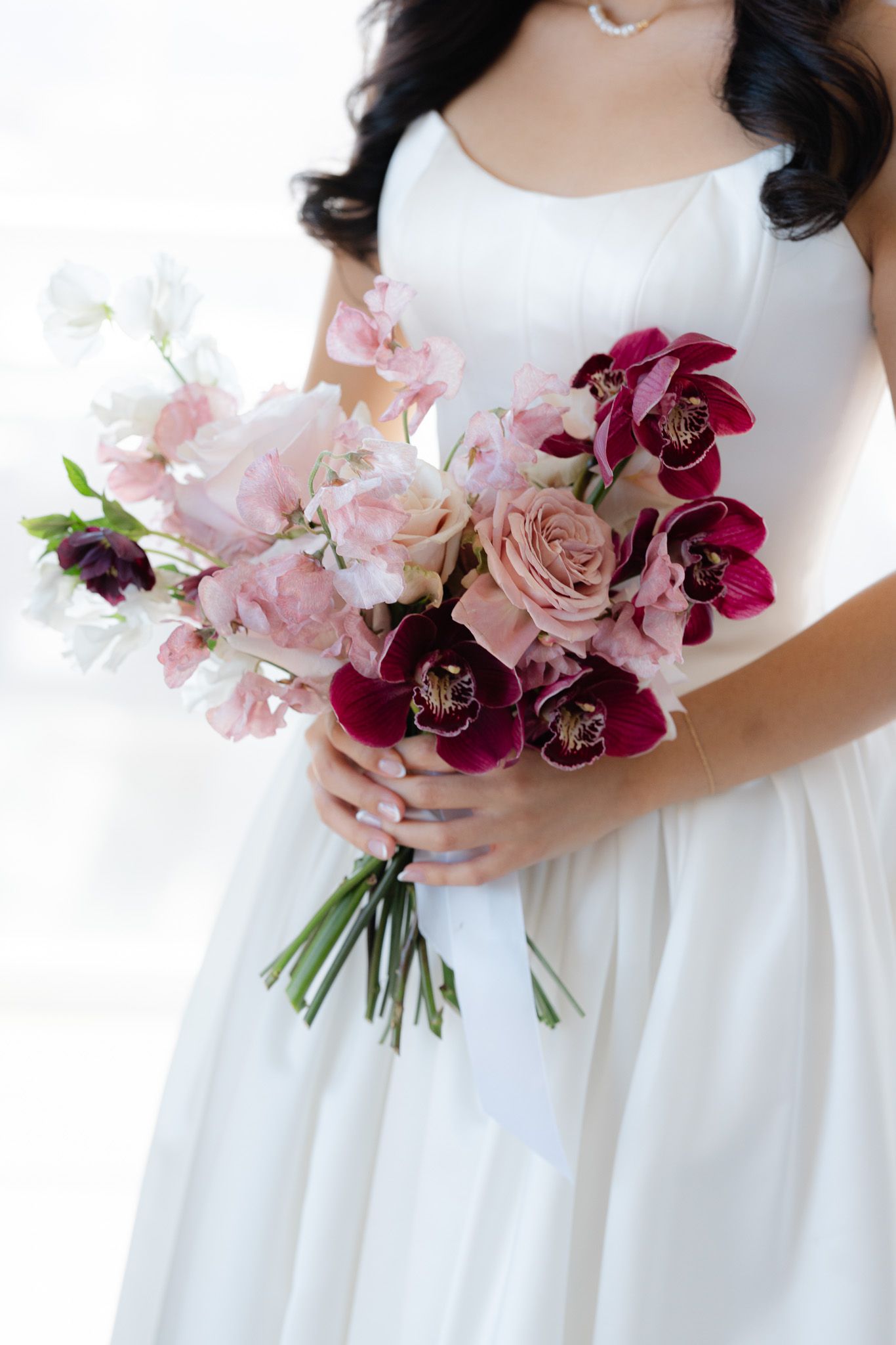 Bride in white dress holding a lush bouquet of pink roses and deep burgundy orchids tied with white ribbon.
