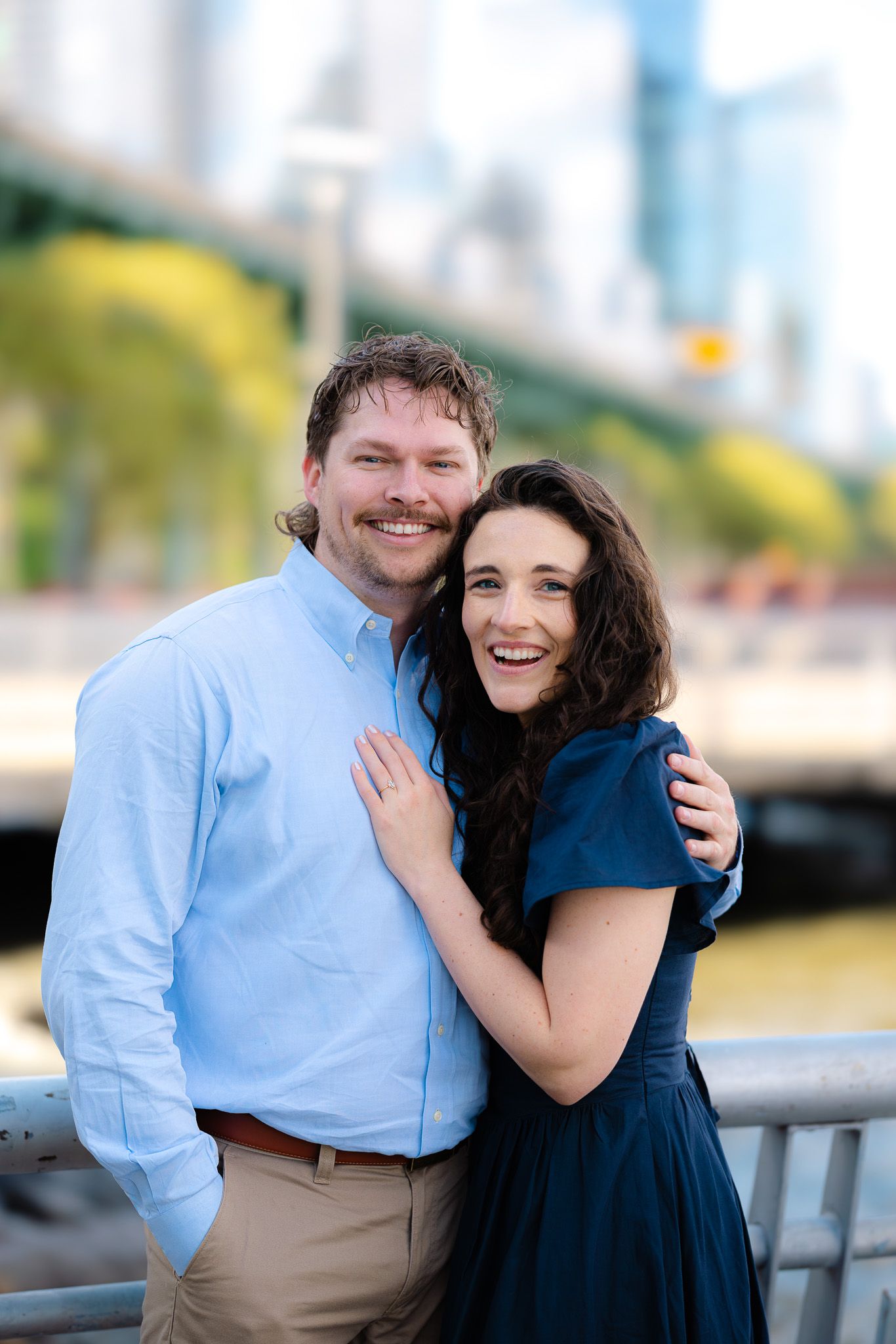 Smiling couple hugging near waterfront railing with NYC bridge and skyline in the background.