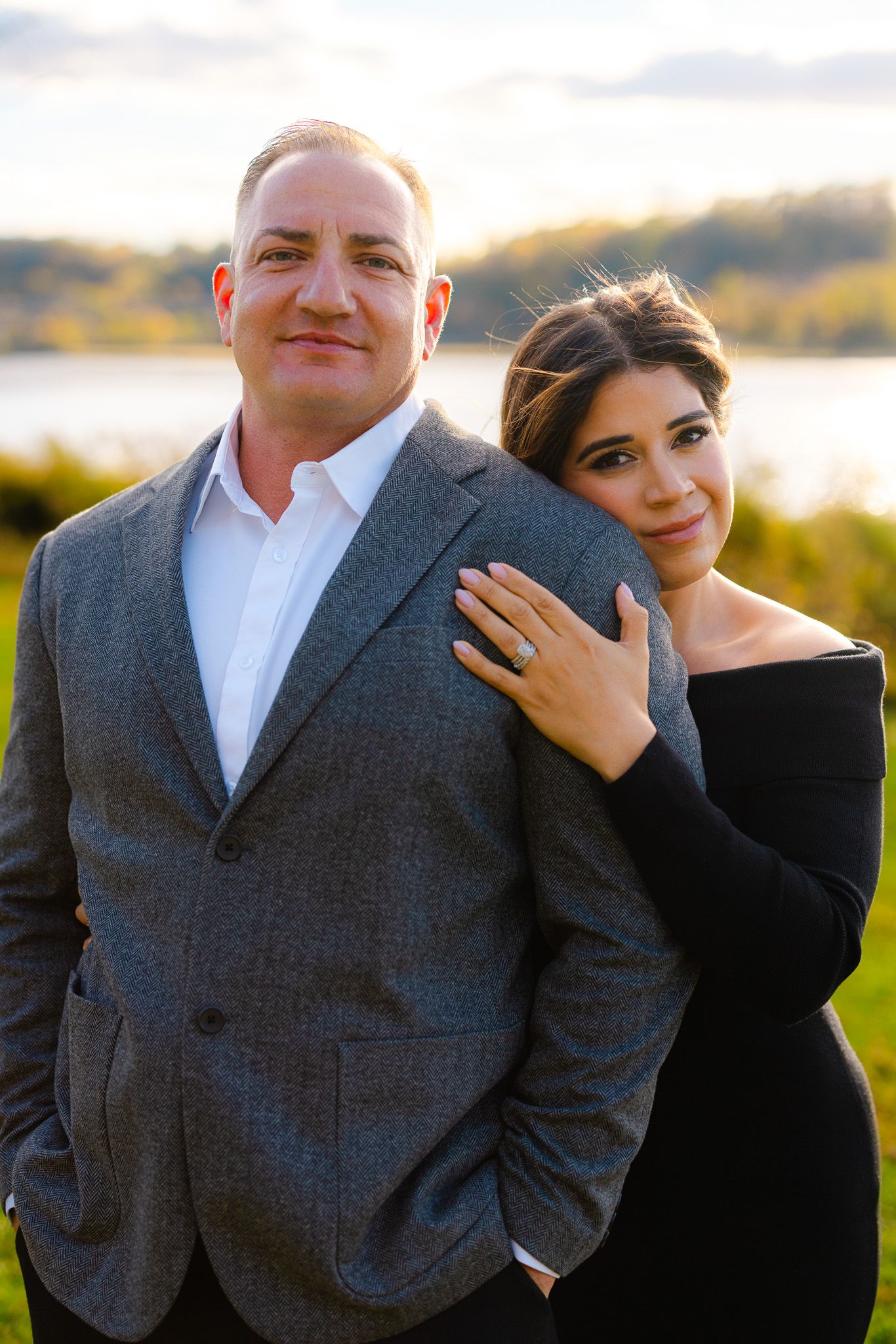Couple poses outdoors at sunset, woman in black dress leans on mans shoulder showing engagement ring.