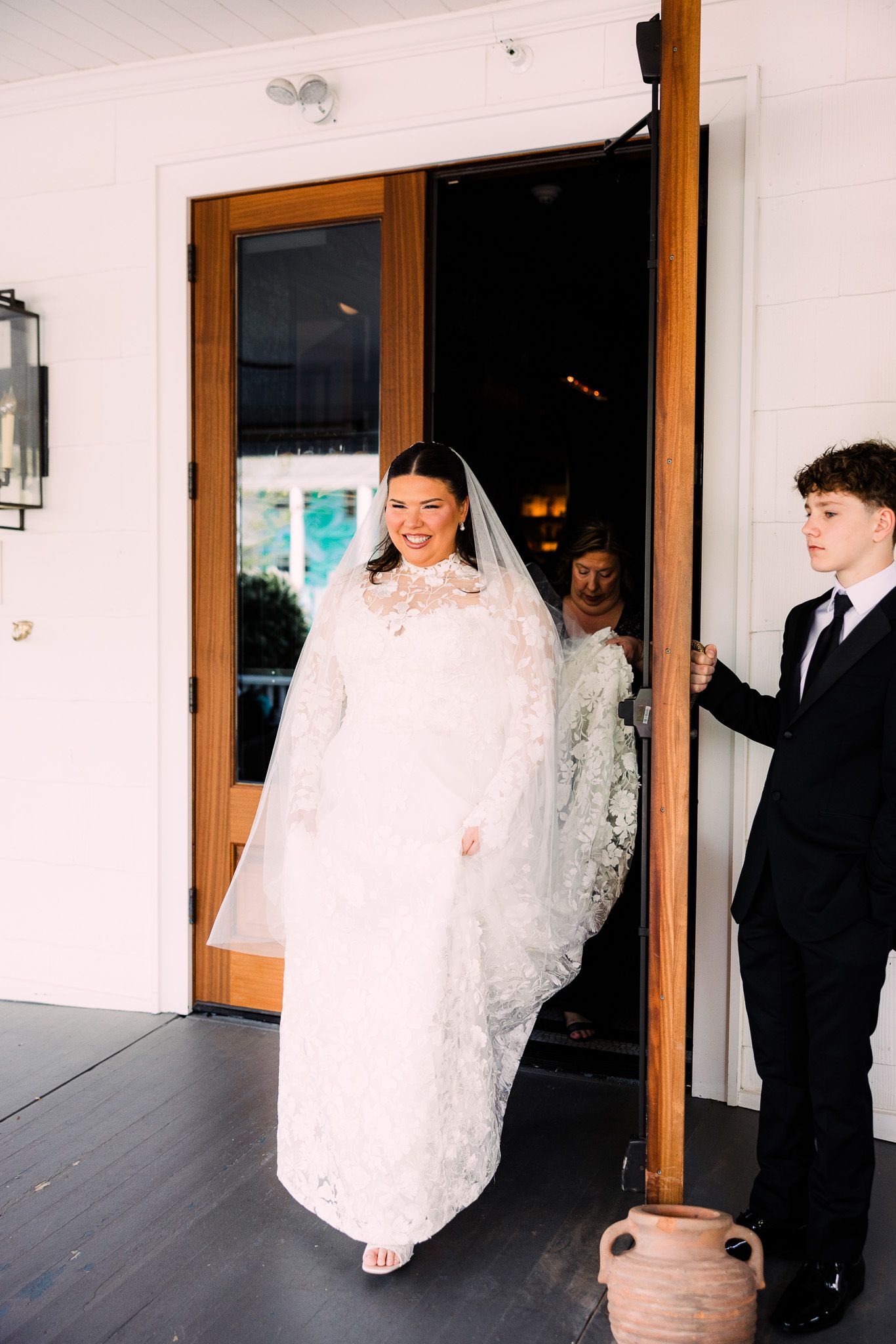 Smiling bride in lace wedding gown with veil walking through wooden doors on wedding day.