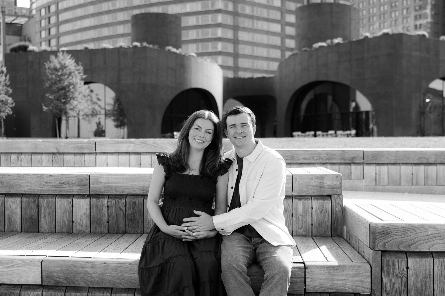 Black and white maternity photo of couple sitting on wooden steps with architectural backdrop.