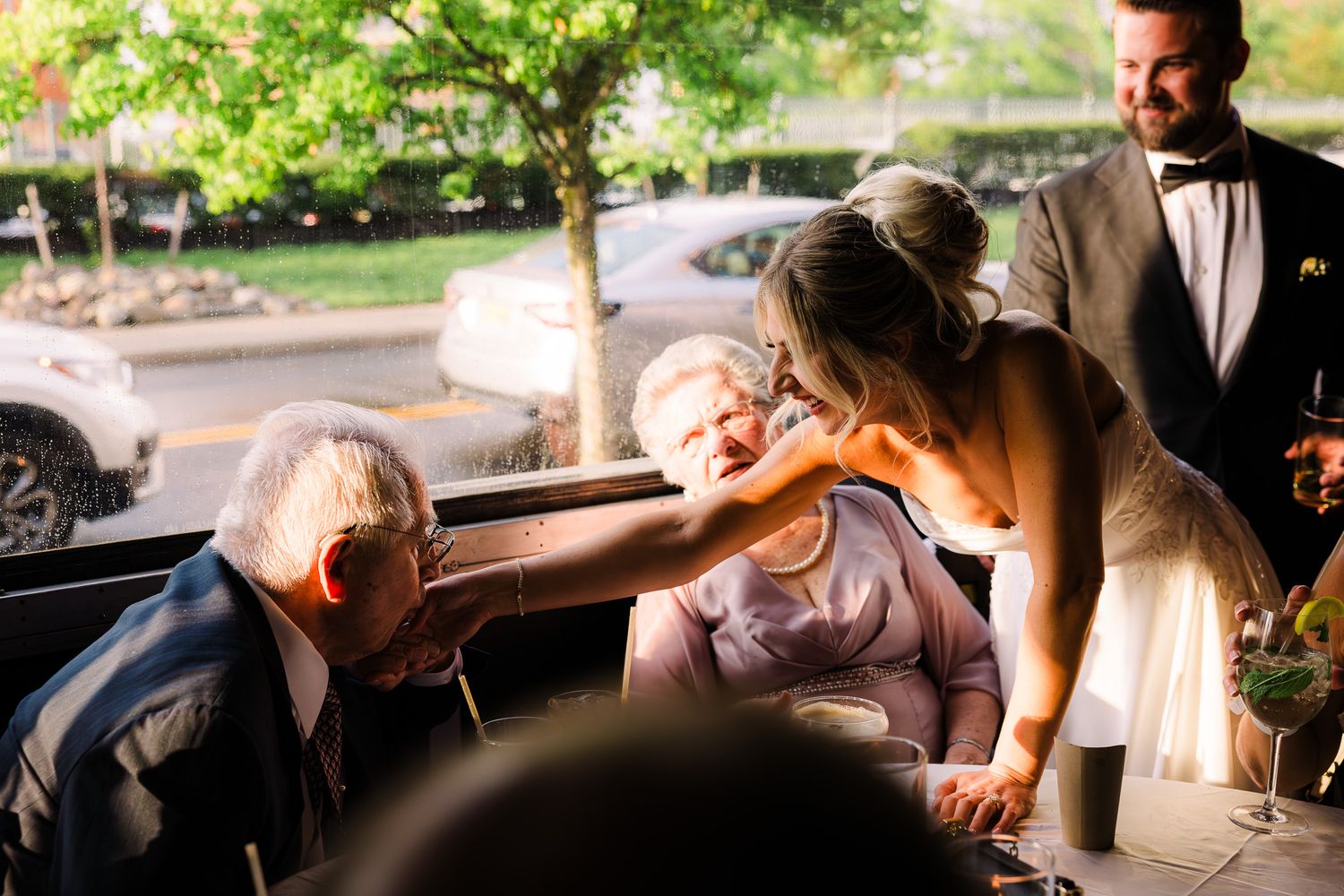 Bride leaning down to greet elderly seated guests at a wedding reception near a window.