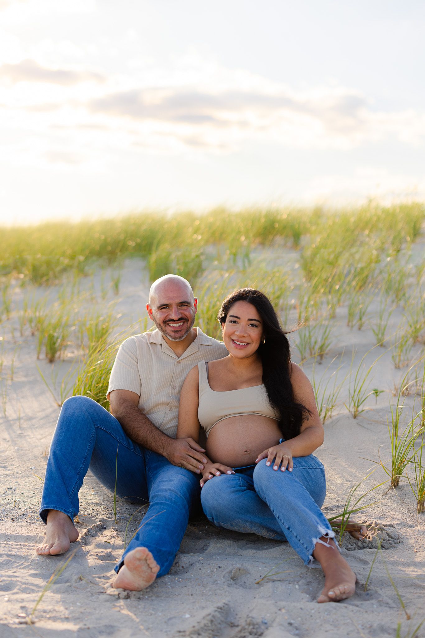 A smiling couple sits on sandy beach dunes, the woman visibly pregnant, both wearing jeans in golden hour light.