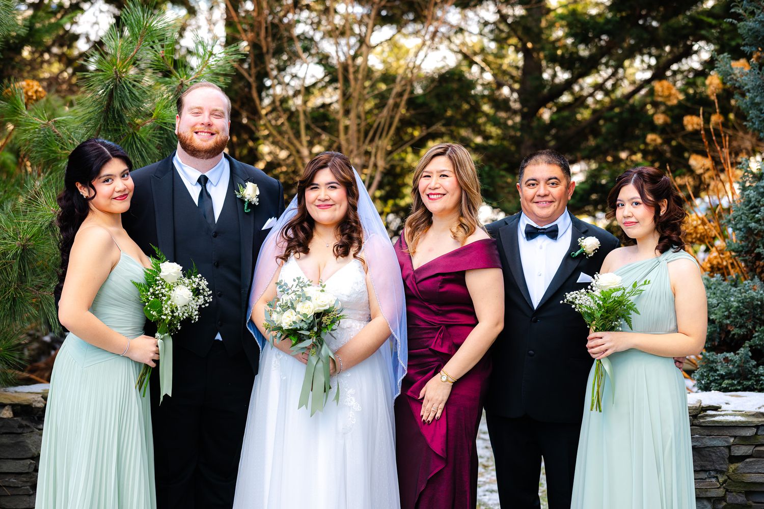Wedding party posing outdoors, bride in white gown with veil, groom in tuxedo, bridesmaids in sage green dresses, holding bouquets.