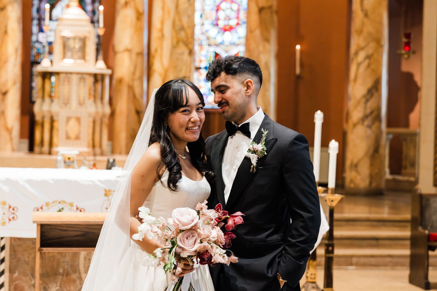 Bride and groom smile at each other inside an ornate church, bride holding a blush and burgundy bouquet.