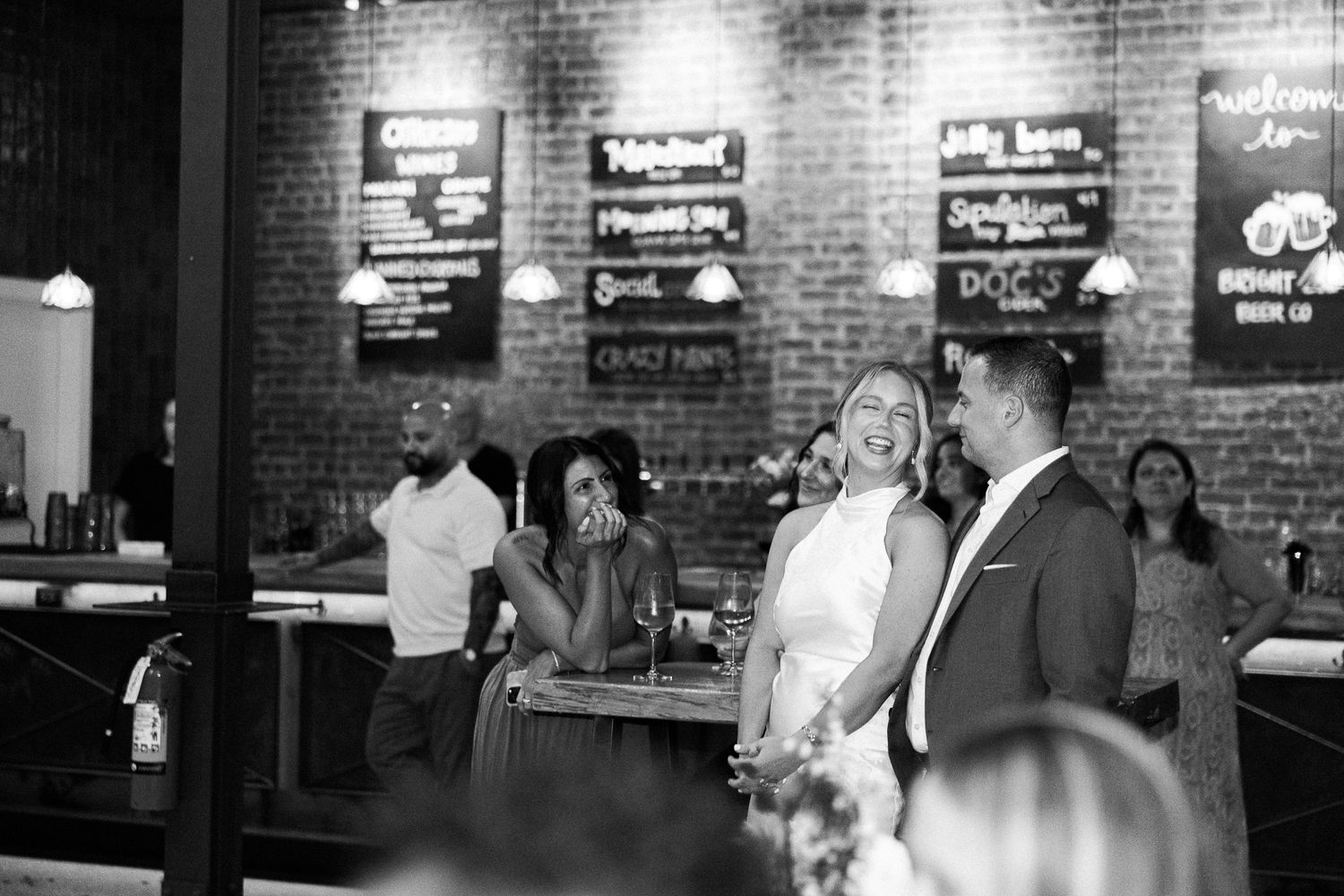 Black and white photo of laughing wedding guests socializing in a rustic brick-walled bar venue during a reception.