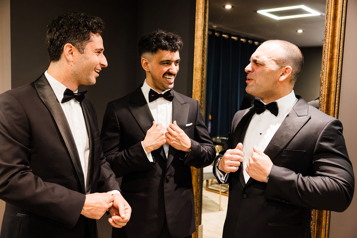Three men in black tuxedos with bow ties laugh and adjust their formal wear before a special event.