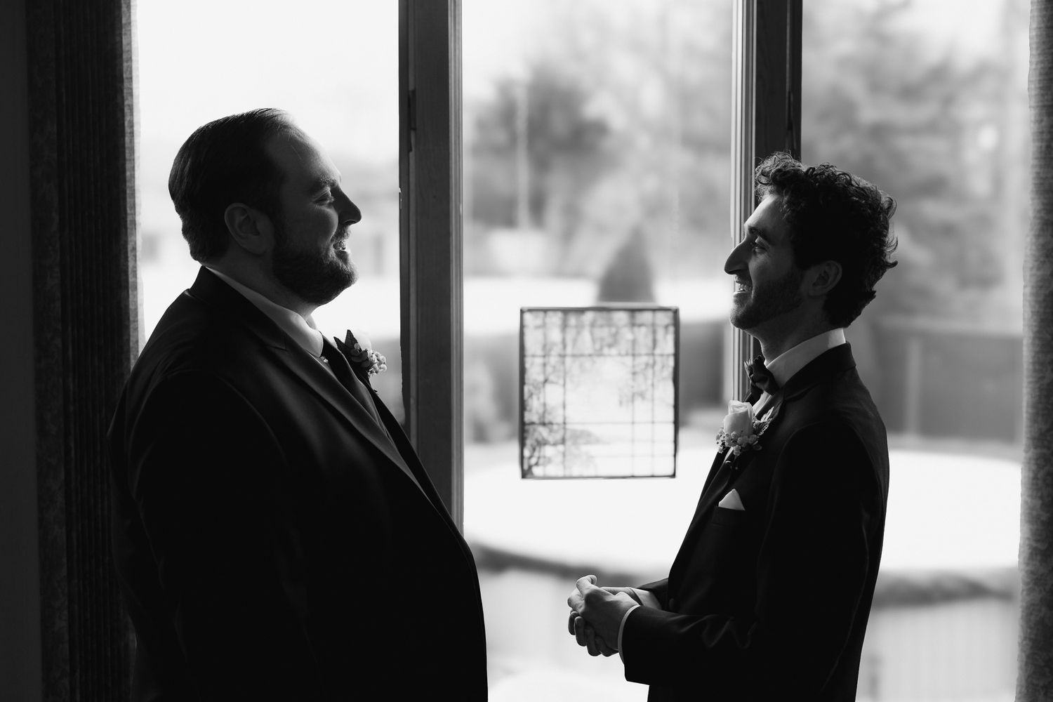 Two grooms in suits share a quiet moment by a window in a black and white wedding photo.