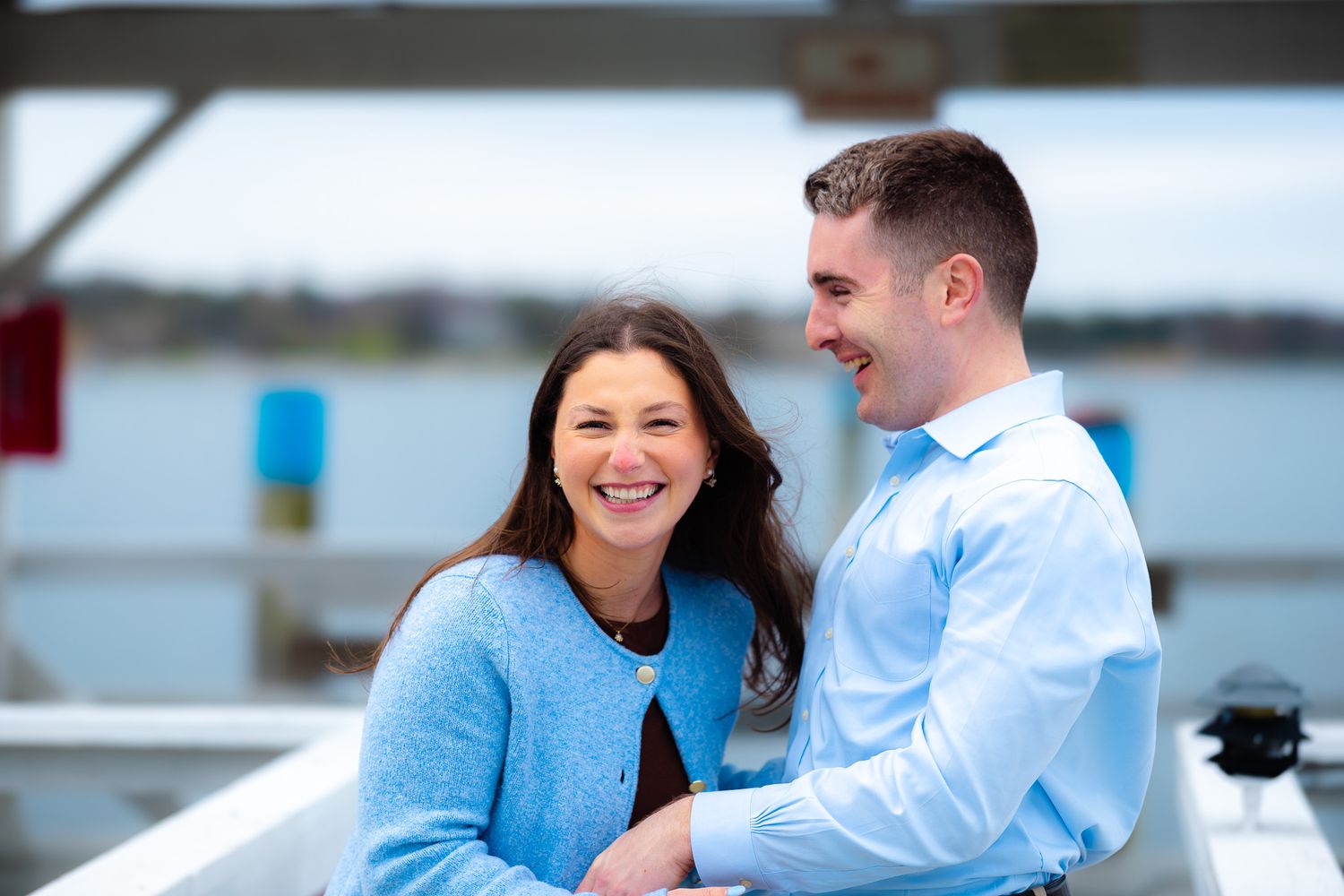 Happy couple laughing together on a waterfront dock, woman in blue cardigan and man in light blue dress shirt.
