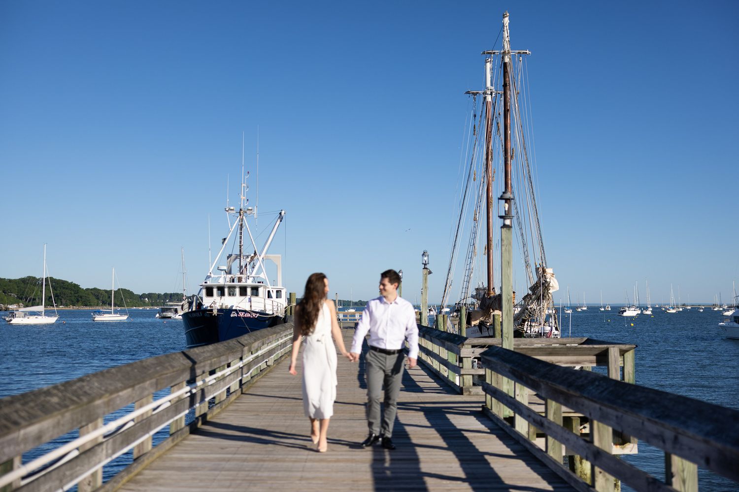 Couple walking hand in hand on a wooden pier by the harbor with sailboats and blue sky.