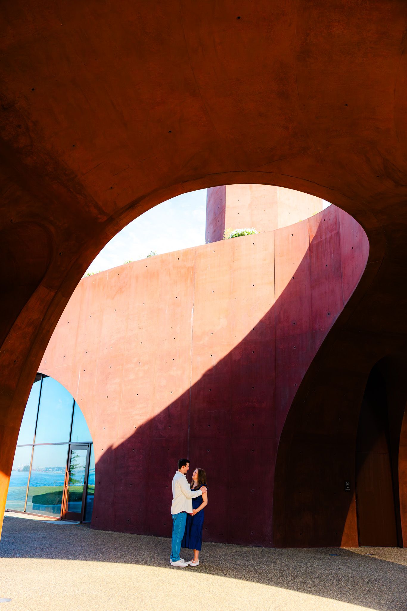 Couple embracing under dramatic curved red concrete architecture with ocean view backdrop.