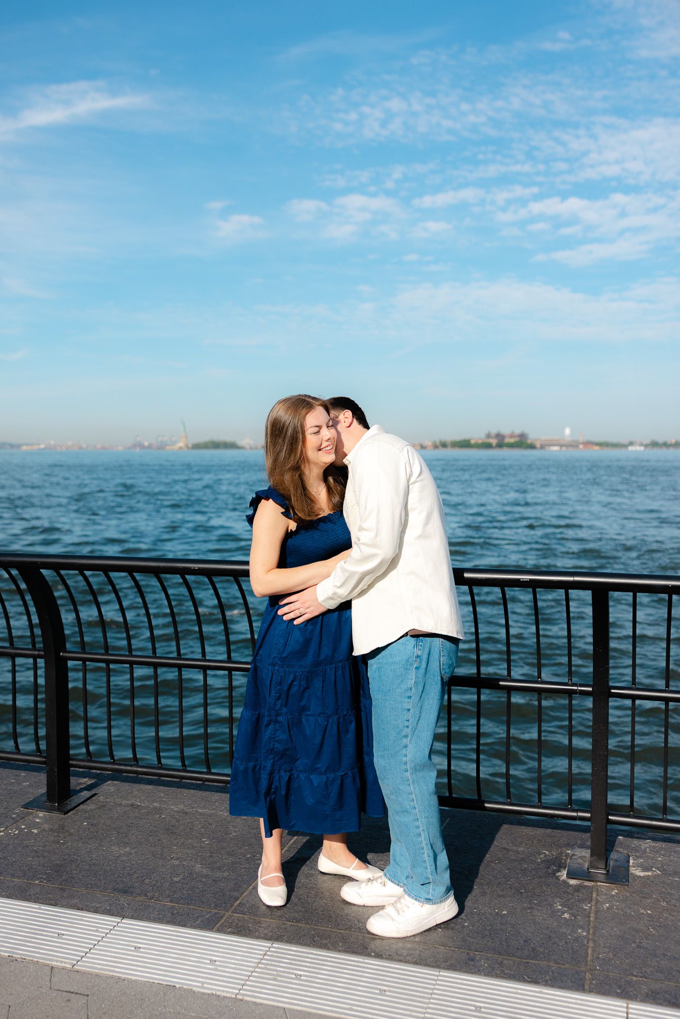 Couple embracing on waterfront promenade with blue sky and harbor views, woman in navy dress, man in white jacket.