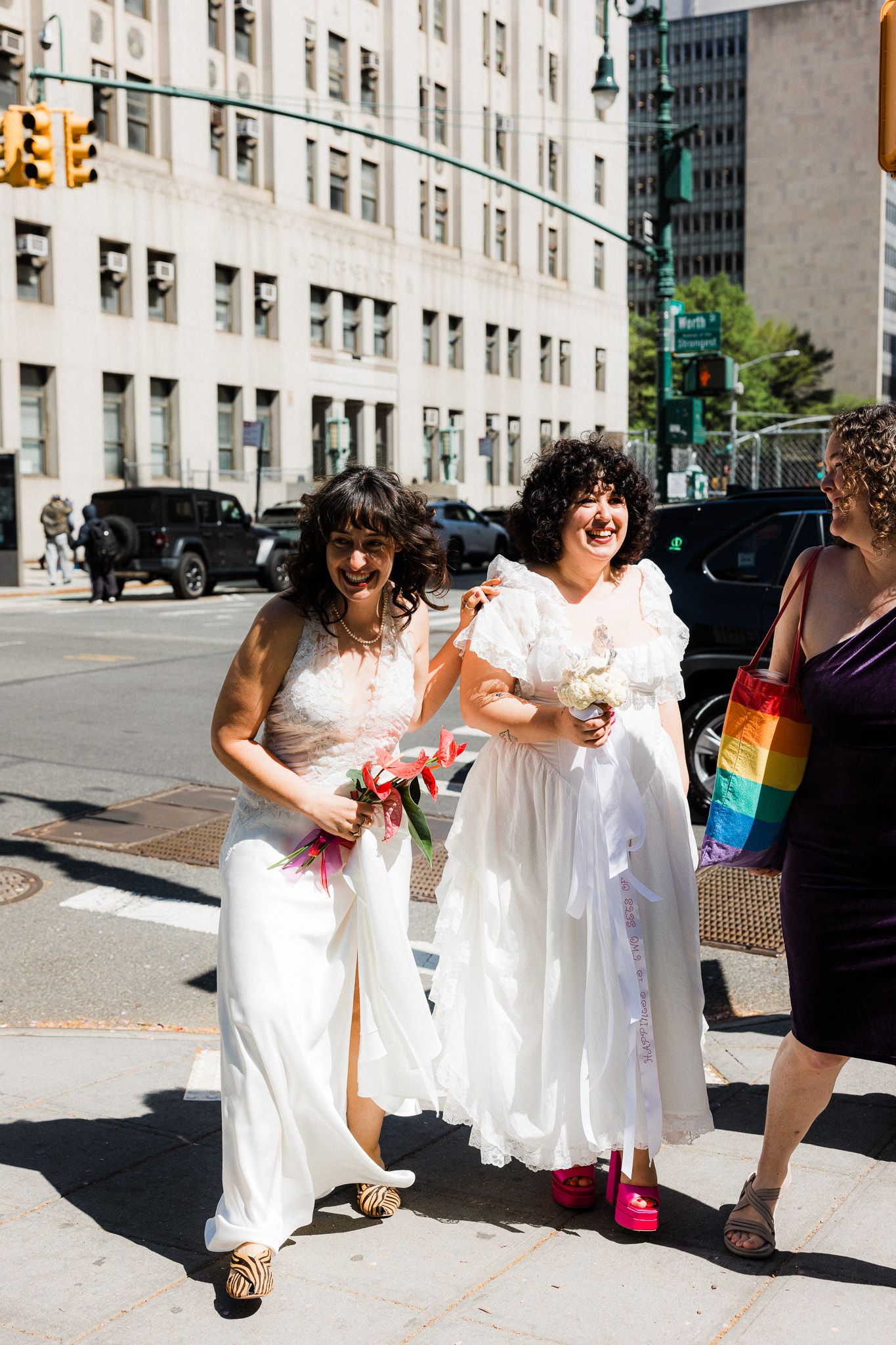 Two brides in white dresses laugh and walk together on a sunny city street holding colorful bouquets.