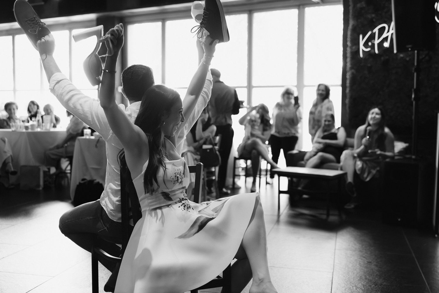 Black and white photo of two people raising shoes in celebration at a lively indoor wedding reception.