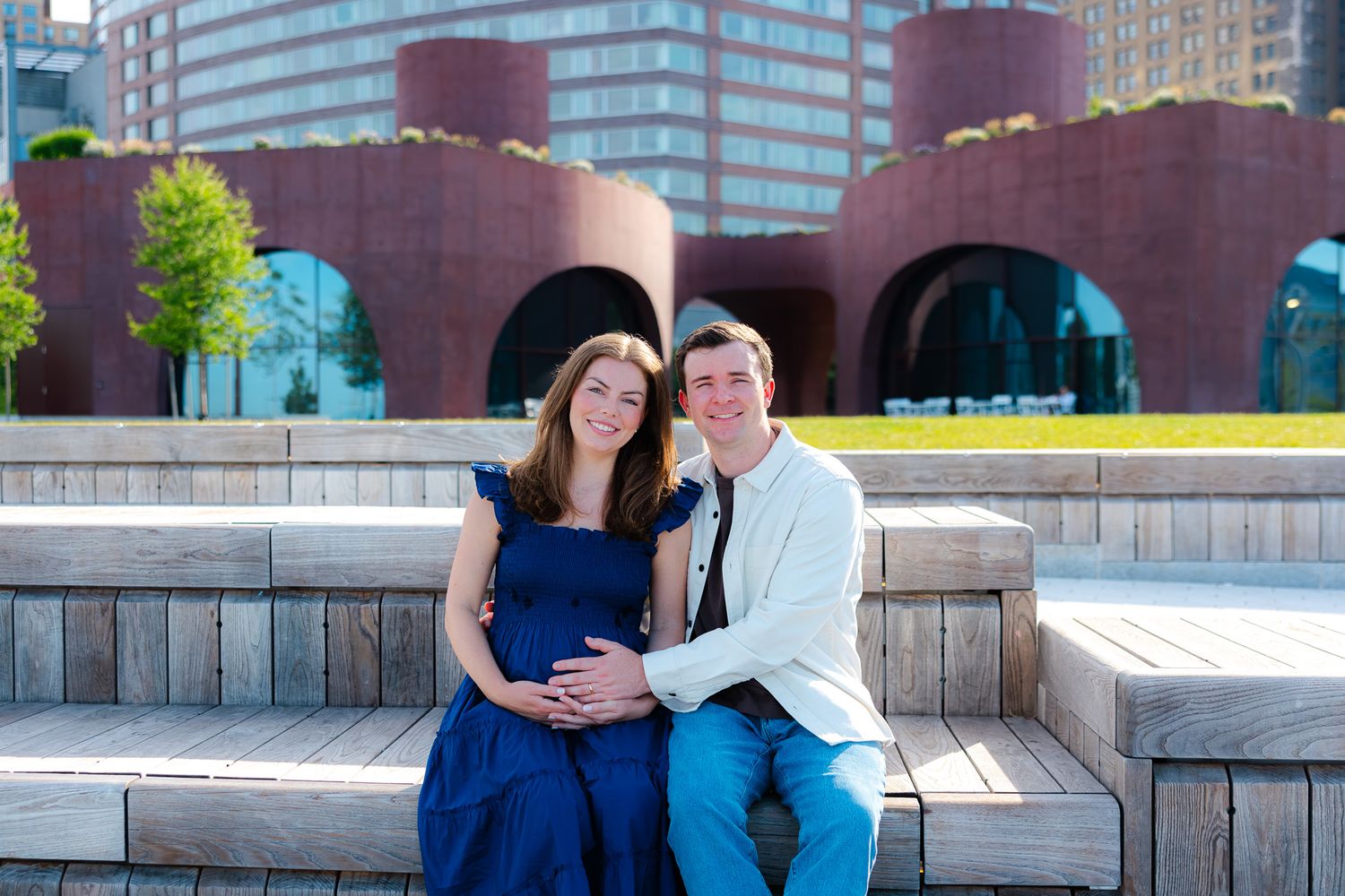 Happy expecting couple posing on wooden steps in blue outfits with red brick building behind them.