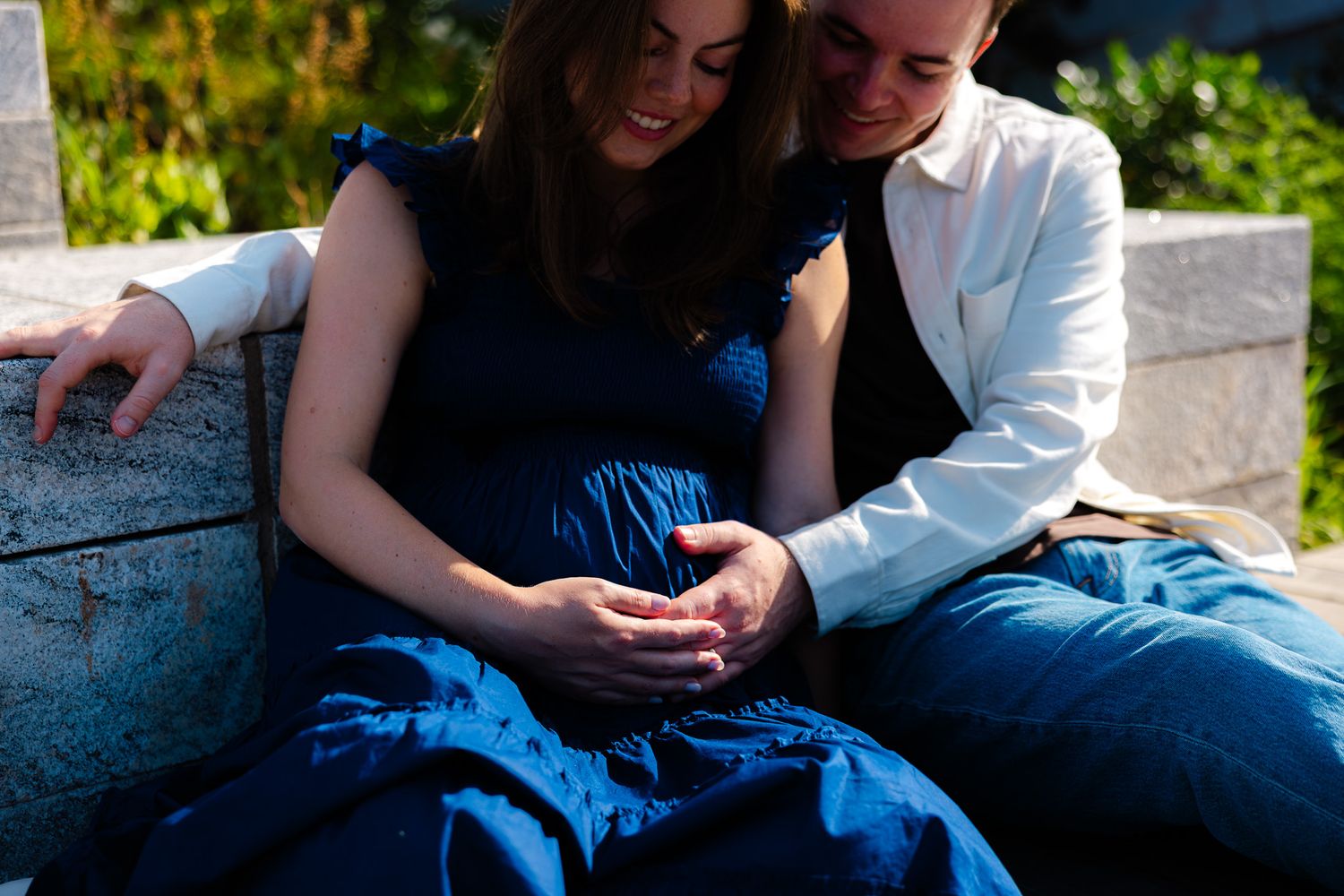 Pregnant couple sitting outdoors, woman in blue dress cradling baby bump with partner in white jacket.