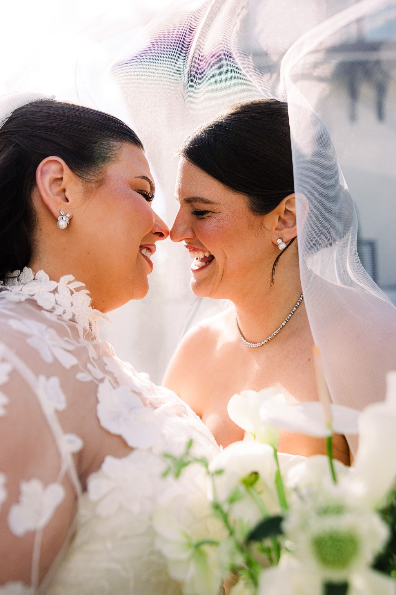 Two brides laughing together under a veil, touching noses in a joyful wedding portrait.