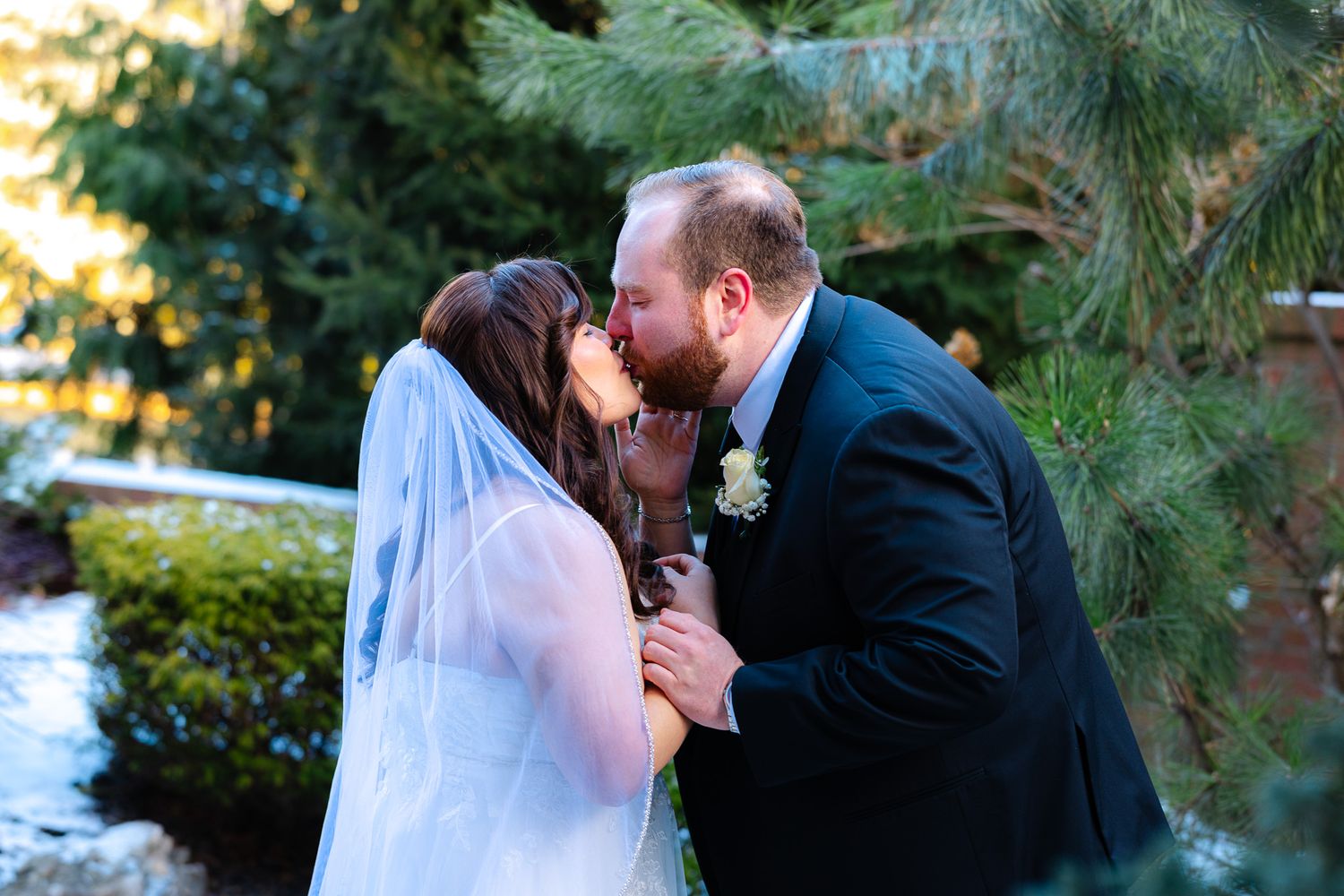 Bride and groom share a romantic first kiss outdoors near pine trees, bride wearing veil and groom in navy suit.