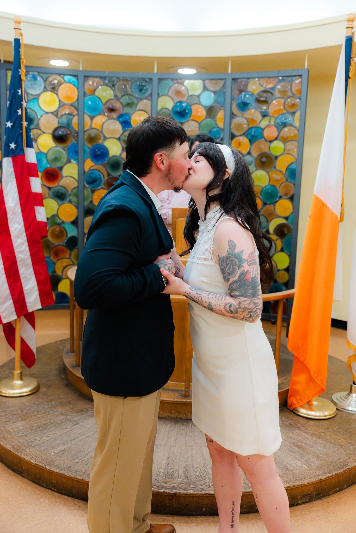 Couple sharing first kiss at city hall wedding ceremony with colorful stained glass backdrop and American flag.