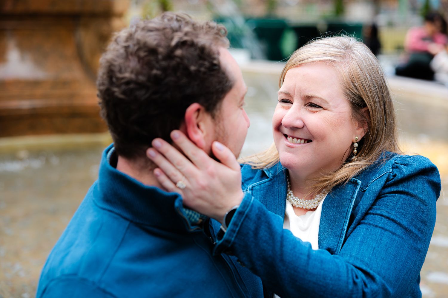 A smiling woman in a blue jacket tenderly touches a man's face outdoors near a fountain.