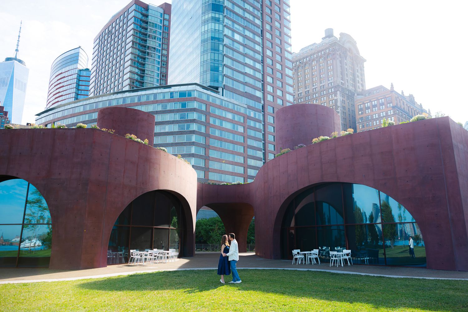 Person standing before large rusty steel arches on green lawn with modern glass skyscrapers in background.