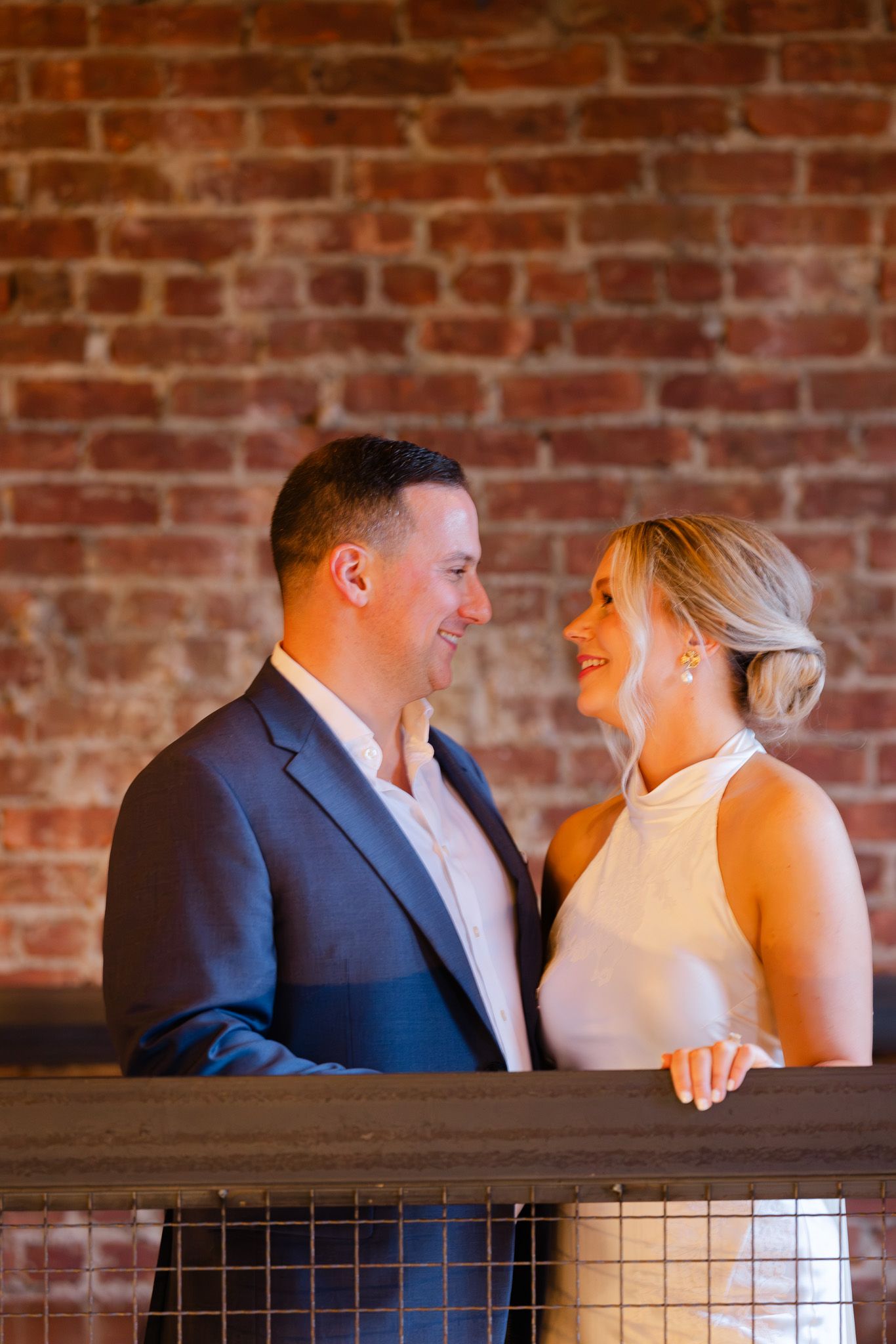 Couple smiling at each other on a balcony with exposed brick wall backdrop, man in navy suit, woman in white dress.