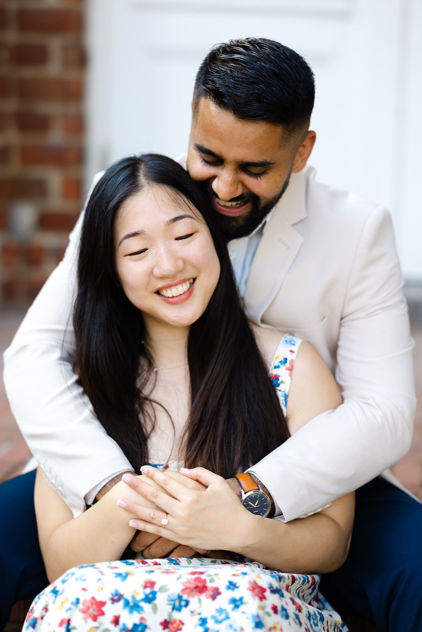 Happy couple embracing outdoors, woman in floral dress smiling, man in white blazer hugging her from behind.