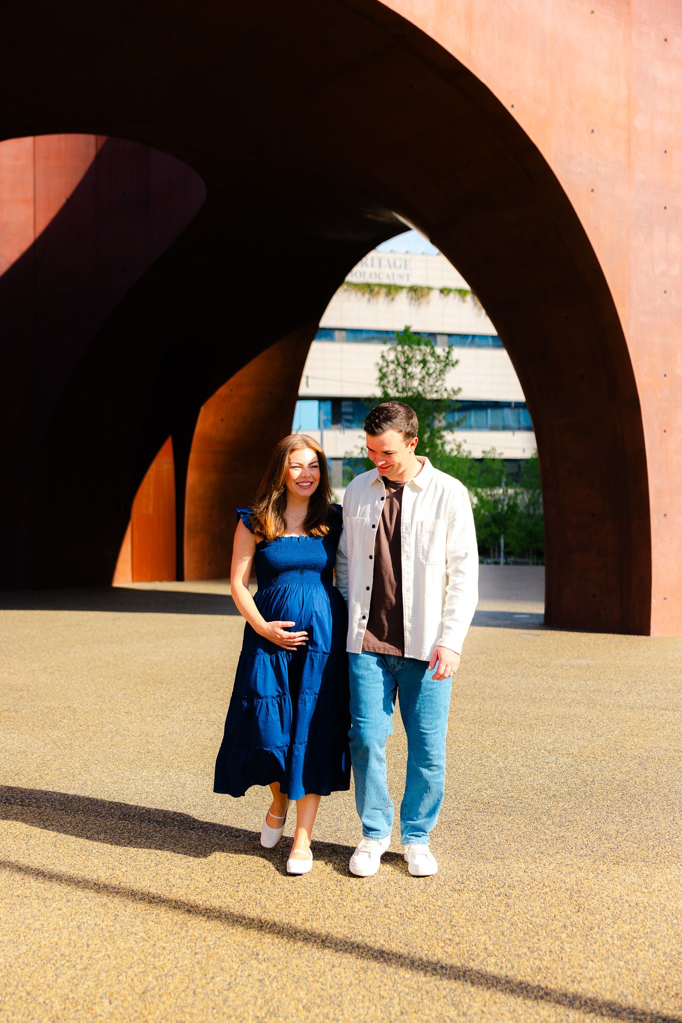 Pregnant woman in blue dress walks with partner under rust-colored arches outdoors.
