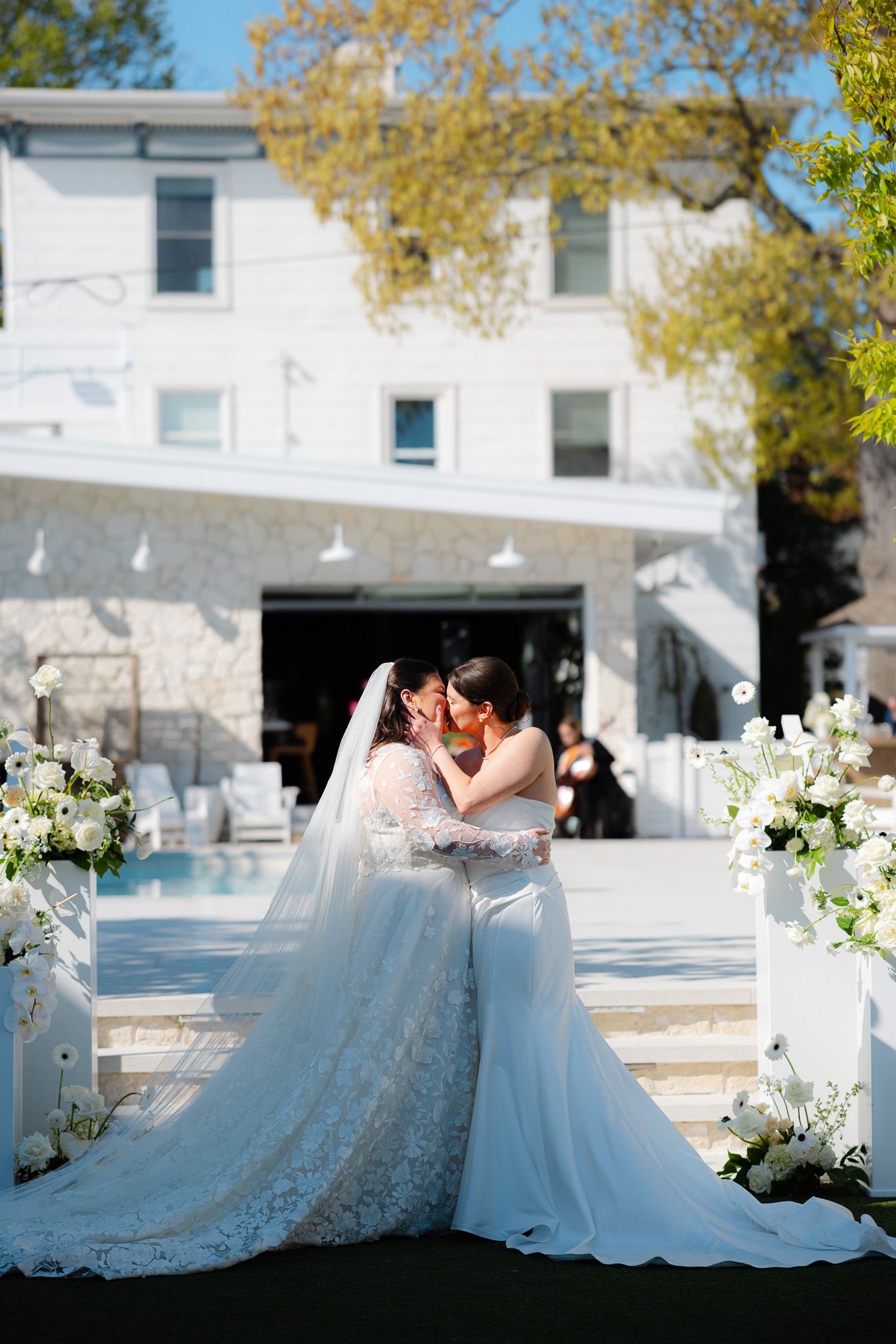 Two brides in white gowns share intimate moment at outdoor wedding ceremony by pool.