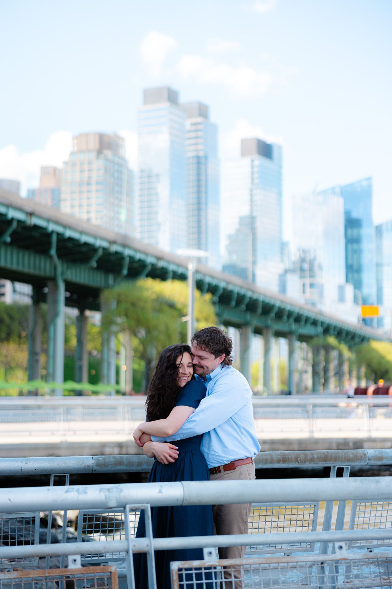 Happy couple embracing at NYC waterfront with Manhattan skyline and elevated highway behind them.