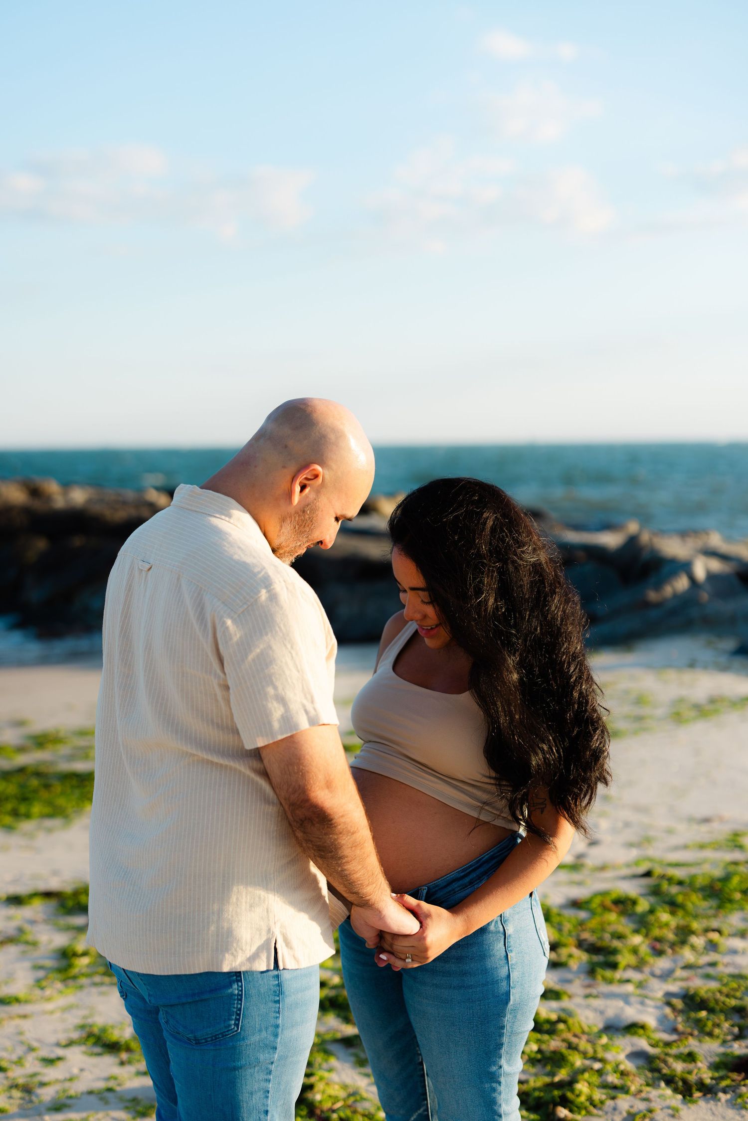 Couple holding hands at beach, pregnant woman showing baby bump, ocean backdrop.