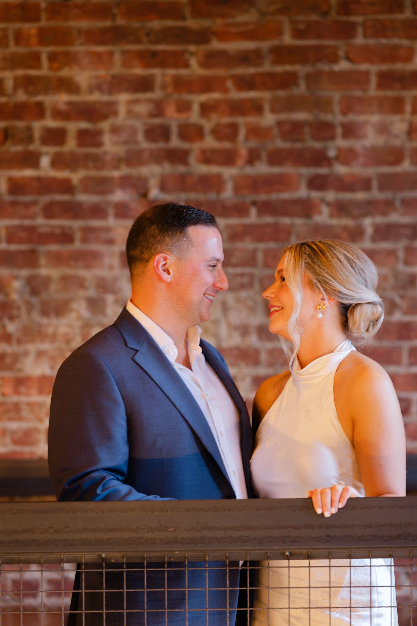 Couple smiling at each other on a balcony with exposed brick wall backdrop, man in navy suit, woman in white dress.