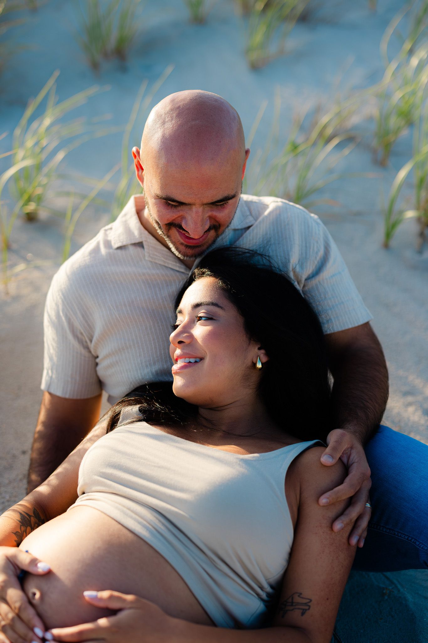 Smiling pregnant woman reclines against her partner on sandy beach dunes, both sharing a tender moment outdoors.