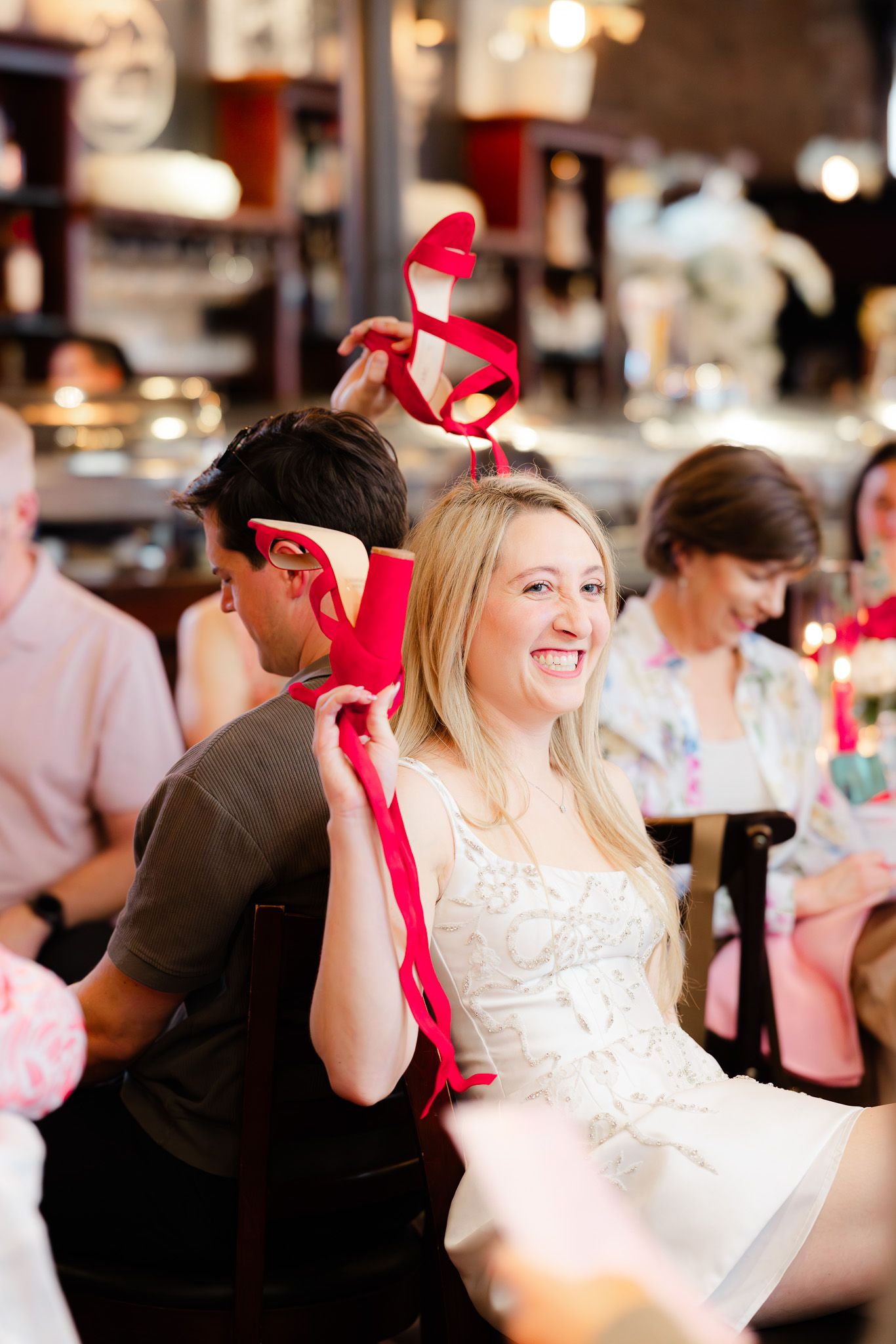Smiling blonde woman holding red high heel shoes at a bridal shower celebration in a restaurant.